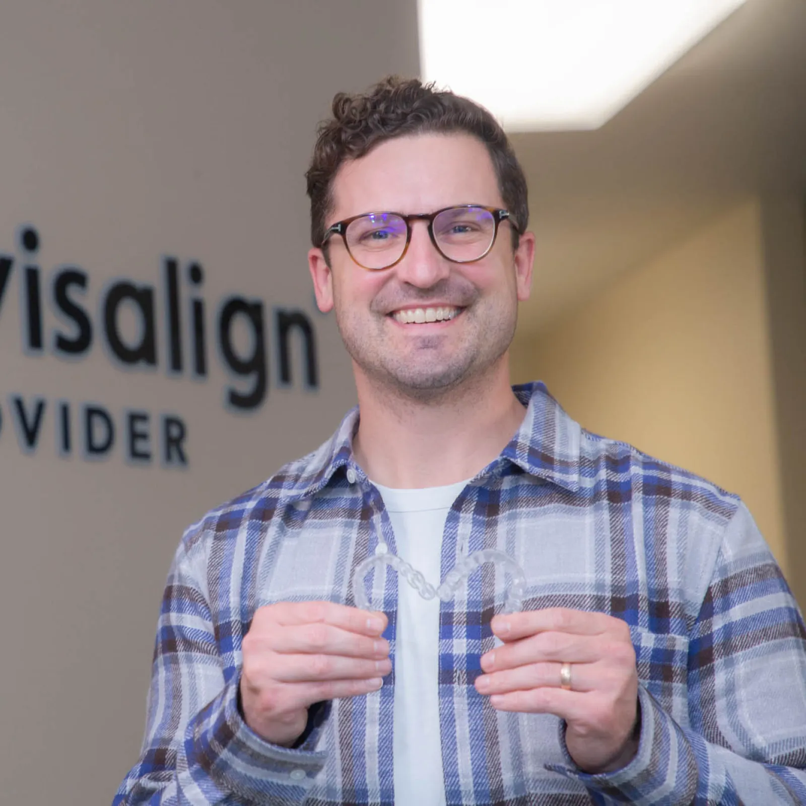 Smiling man holding clear Invisalign aligner in dental office with Invisalign Provider sign in background