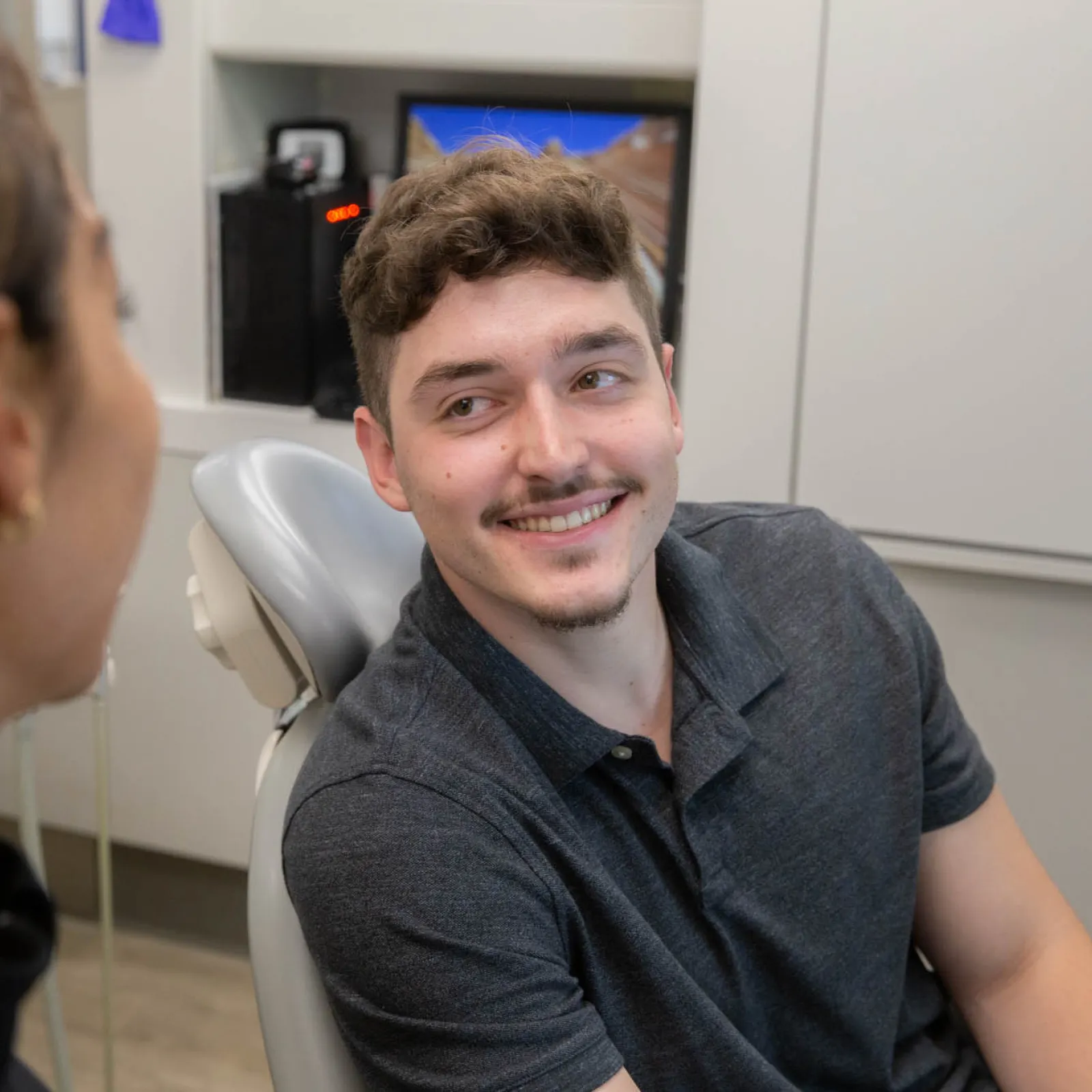 Smiling young man sitting in a dental chair talking to a female dental professional in a clinic.