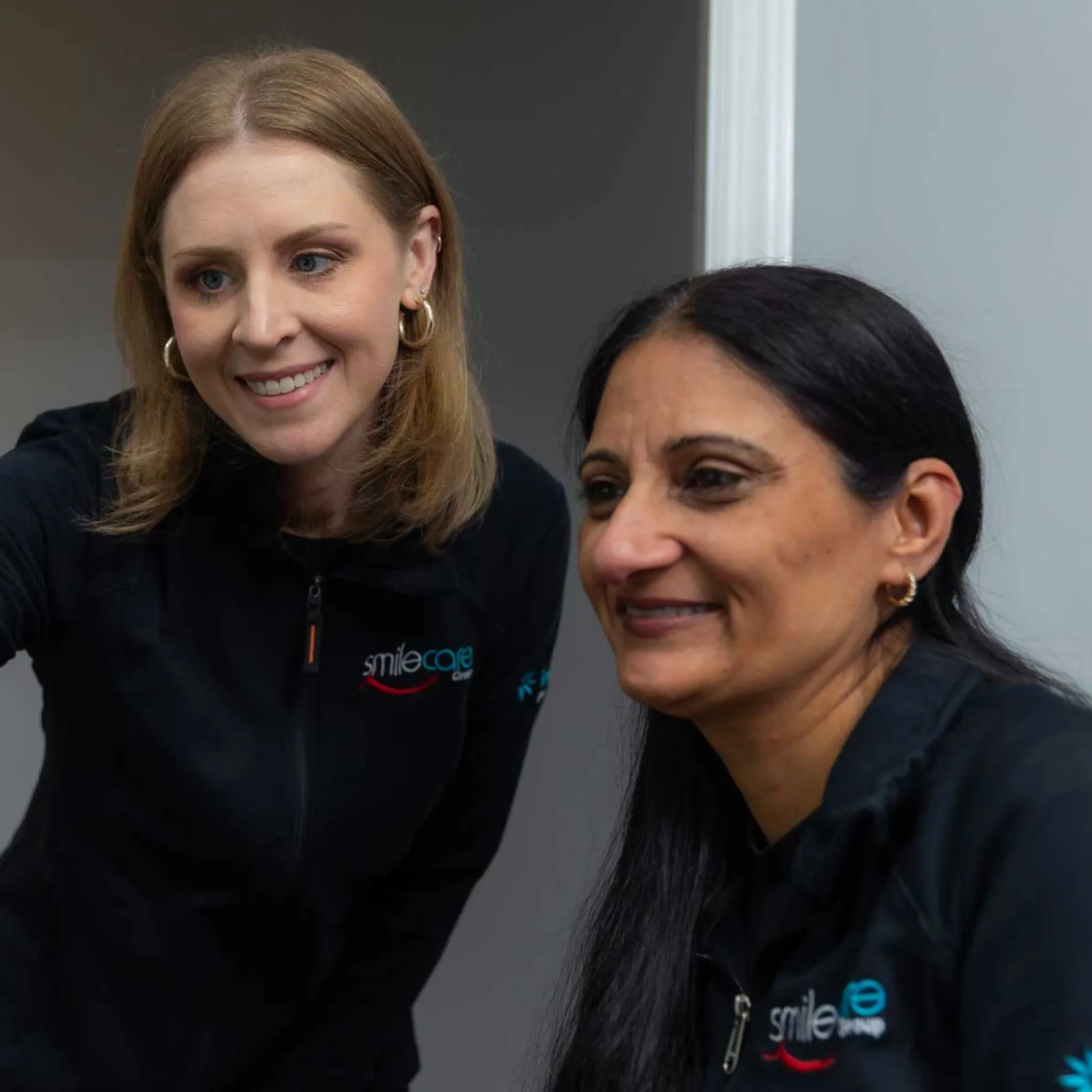 Two female dental professionals in Smile Care uniforms discussing patient information on a computer screen.