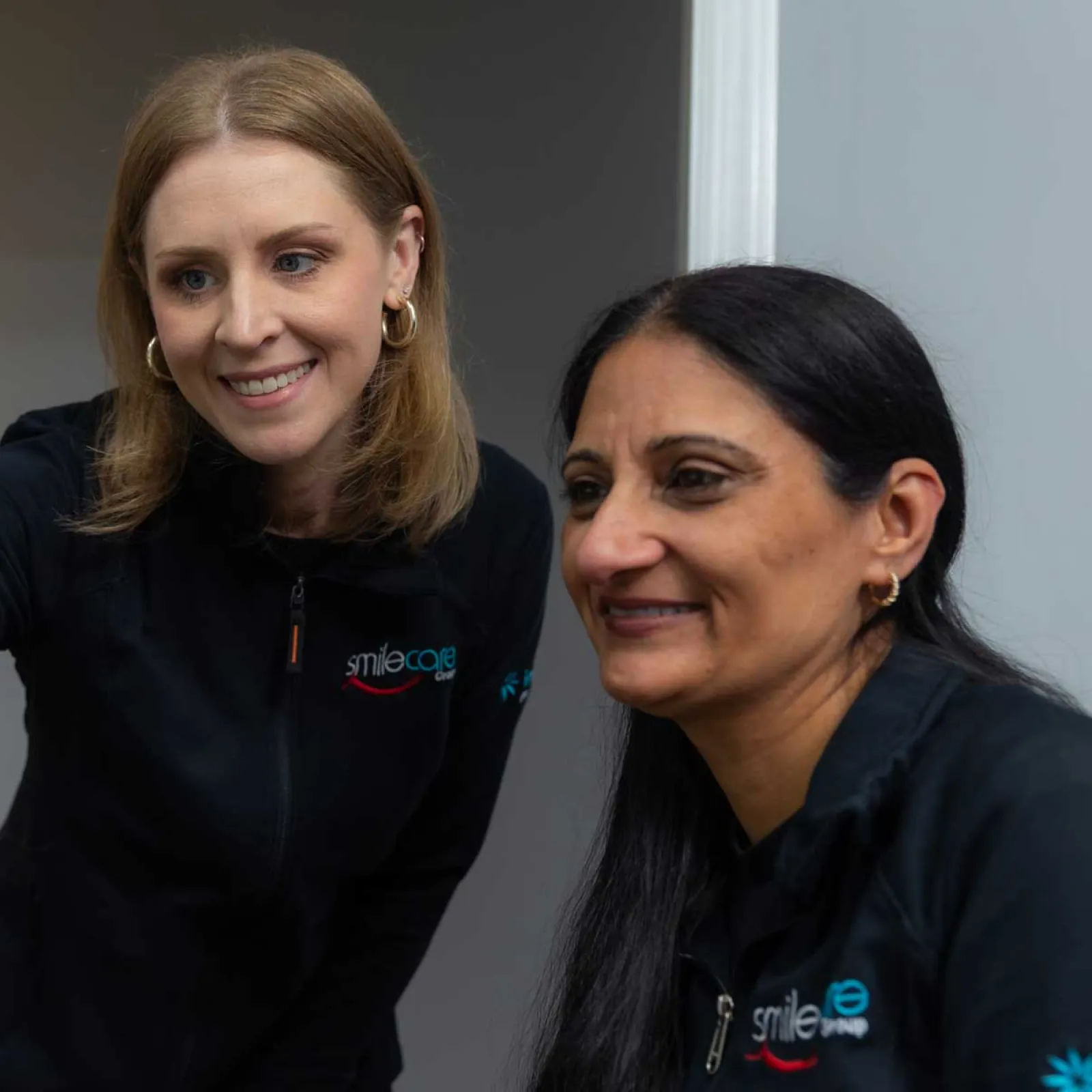 Two female dental professionals in Smile Care uniforms discussing patient information on a computer screen.