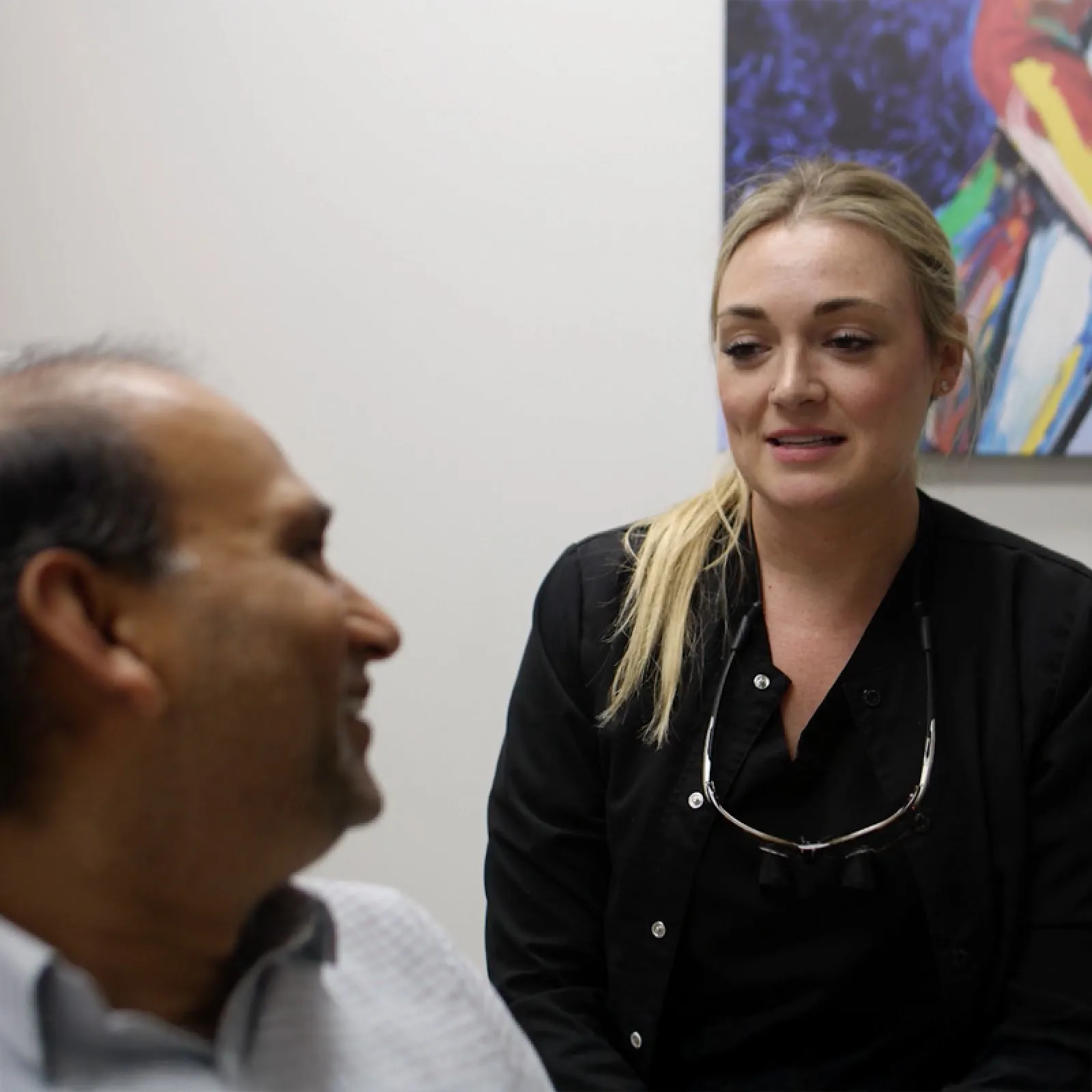 Dentist consulting with a male patient in a modern clinic, wearing black scrubs and dental loupes.