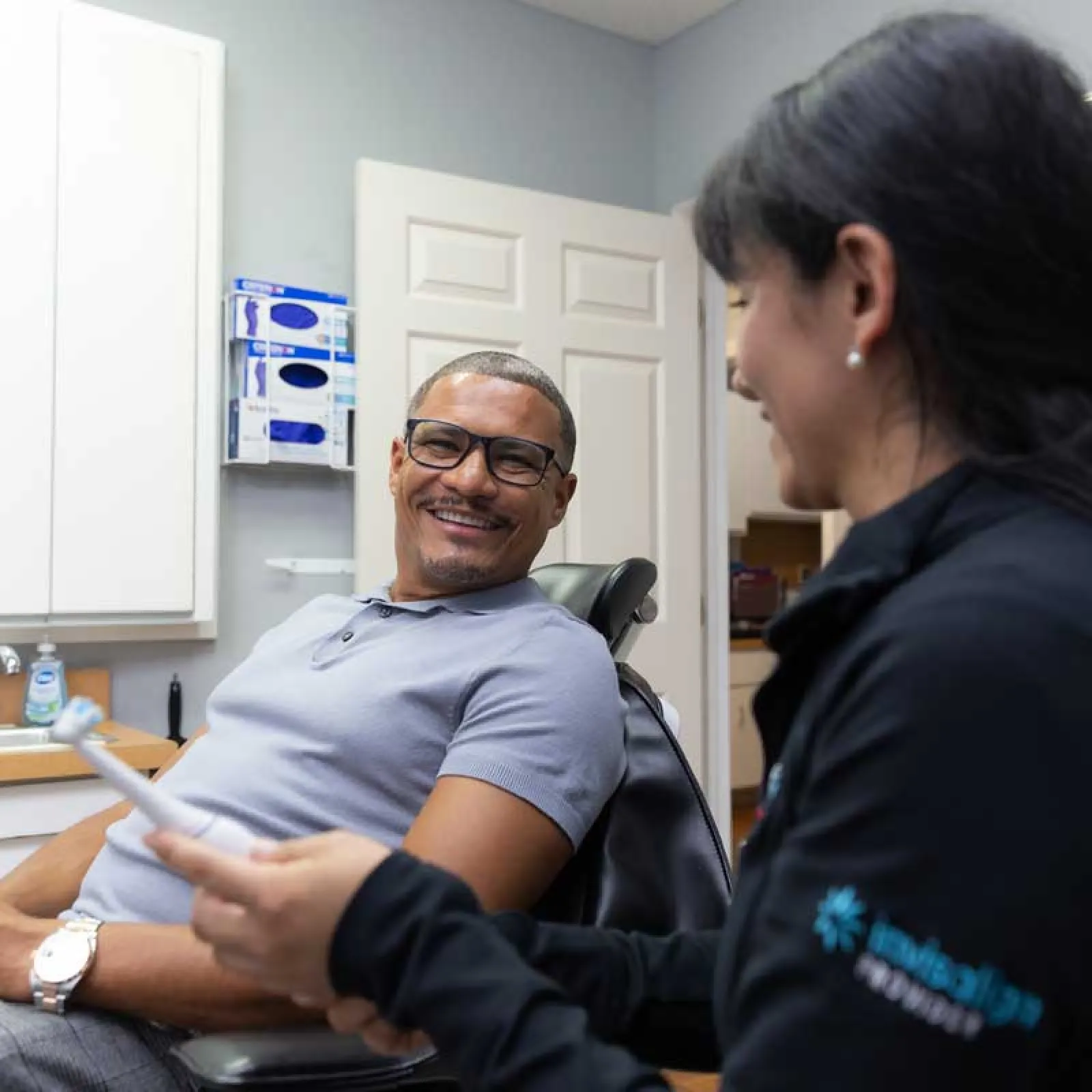 Smiling man sits in dental chair talking to female dental professional holding a dental tool in clinic.