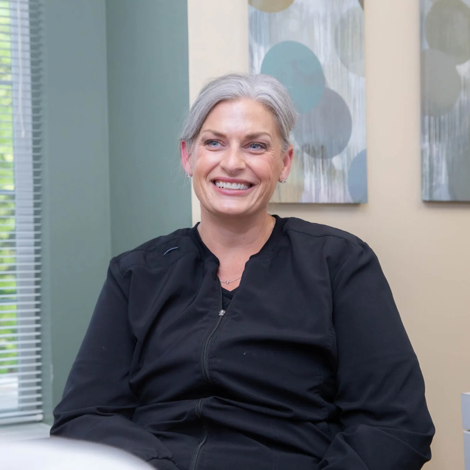 Smiling middle-aged woman with gray hair wearing black jacket sitting indoors in a bright room.