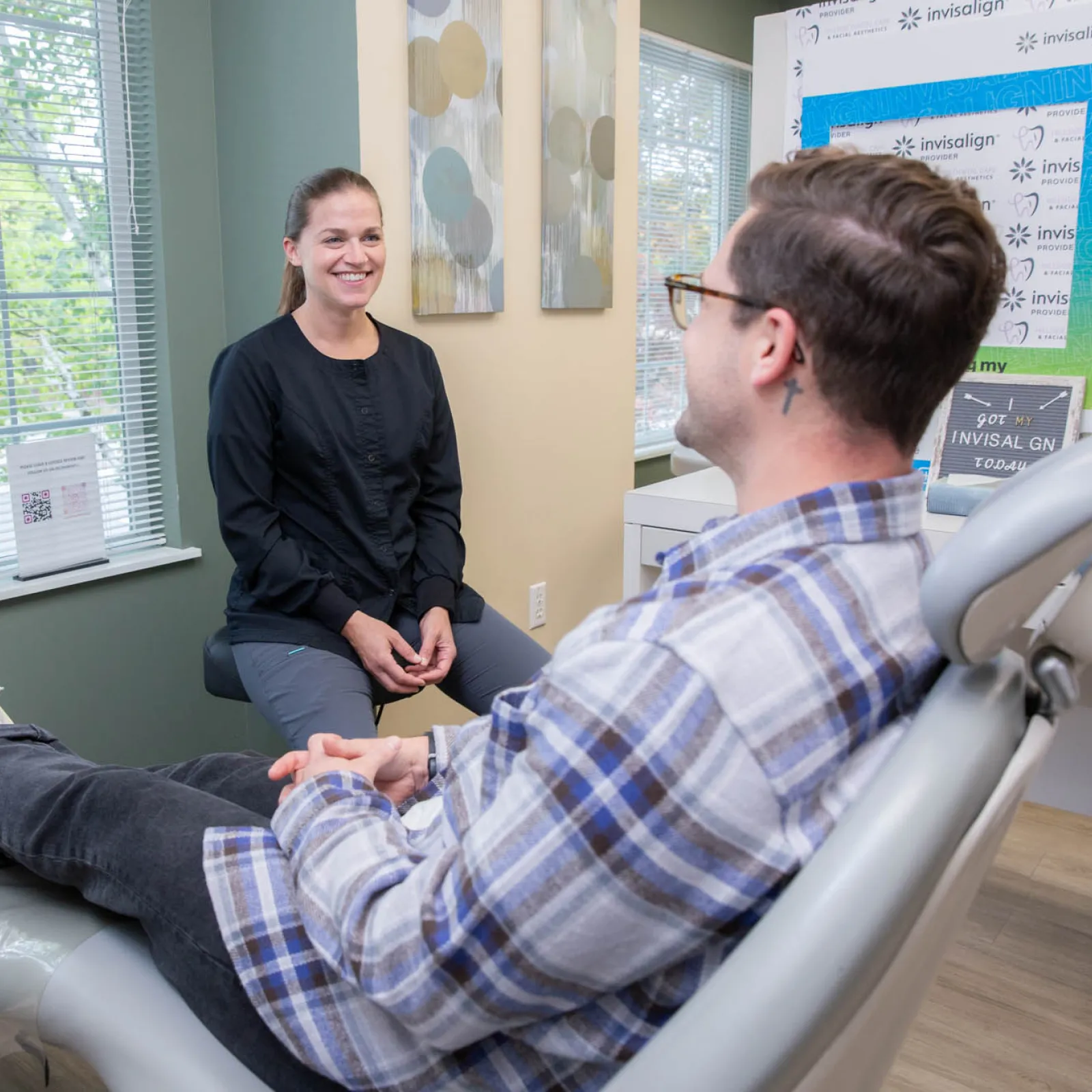 Patient sitting in dental chair talking with smiling dental professional in a bright office with window and Invisalign signs.