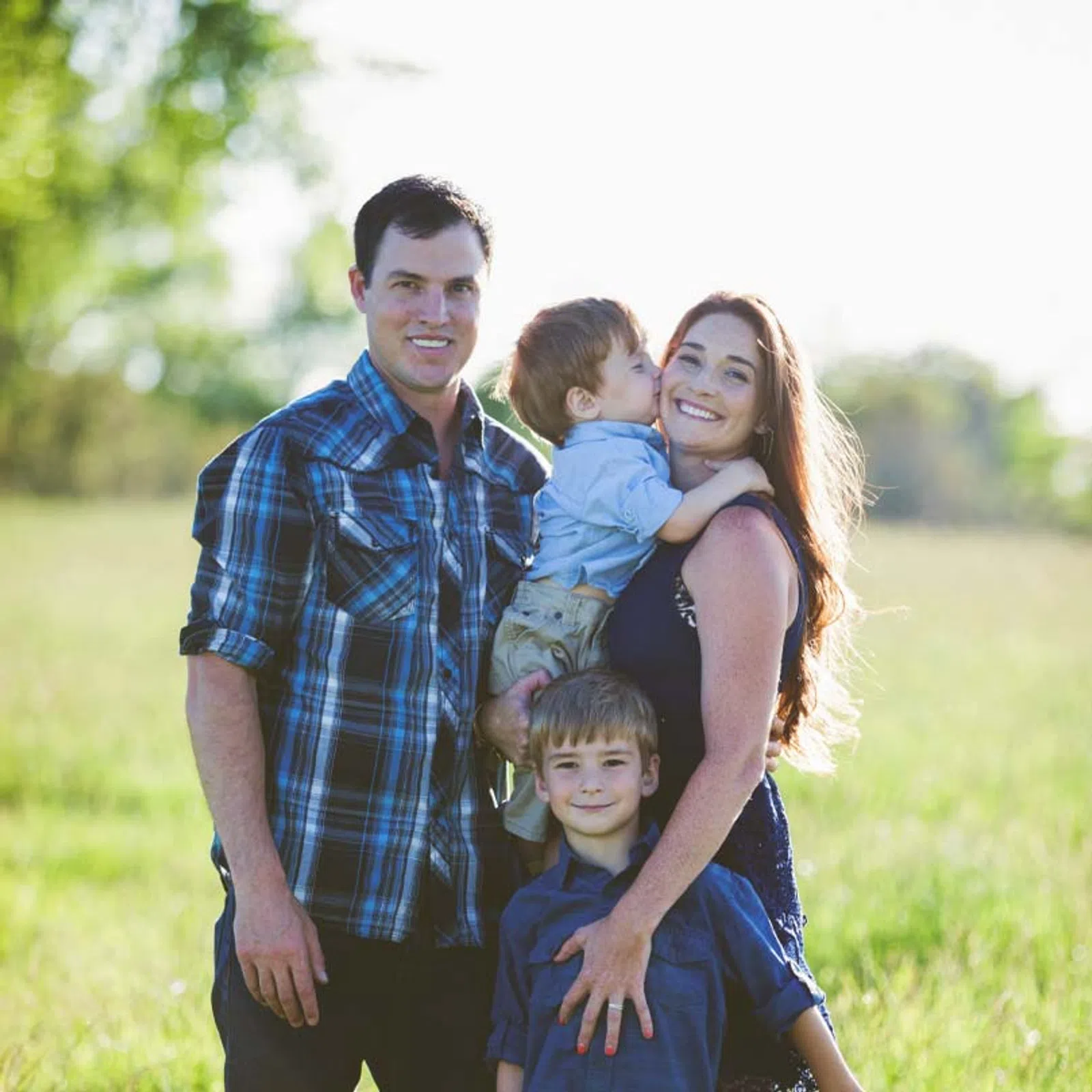 Smiling family of four with two young boys posing outdoors in a sunlit grassy field on a clear day