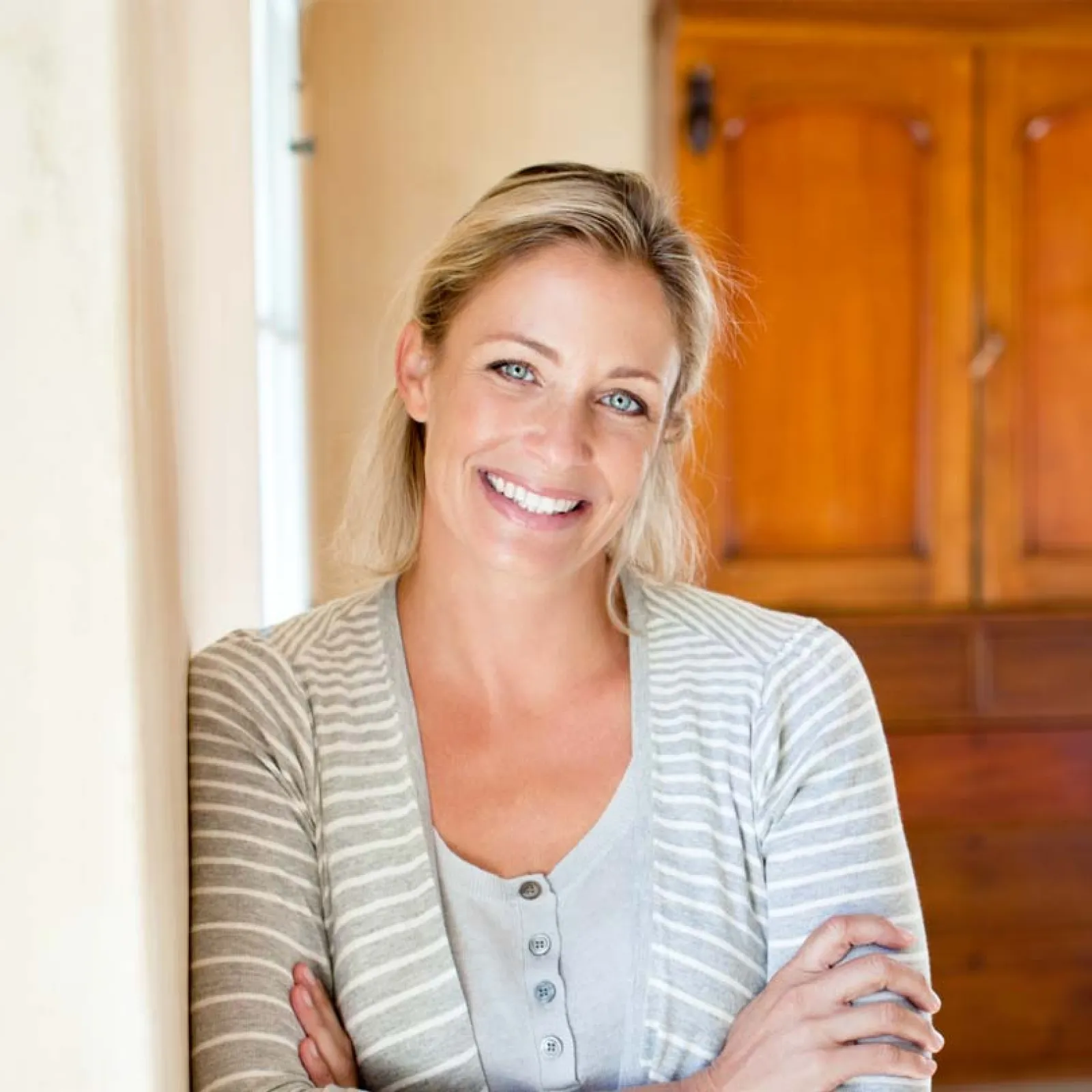Smiling blonde woman in light gray striped cardigan leaning against wall with wooden cabinet in background
