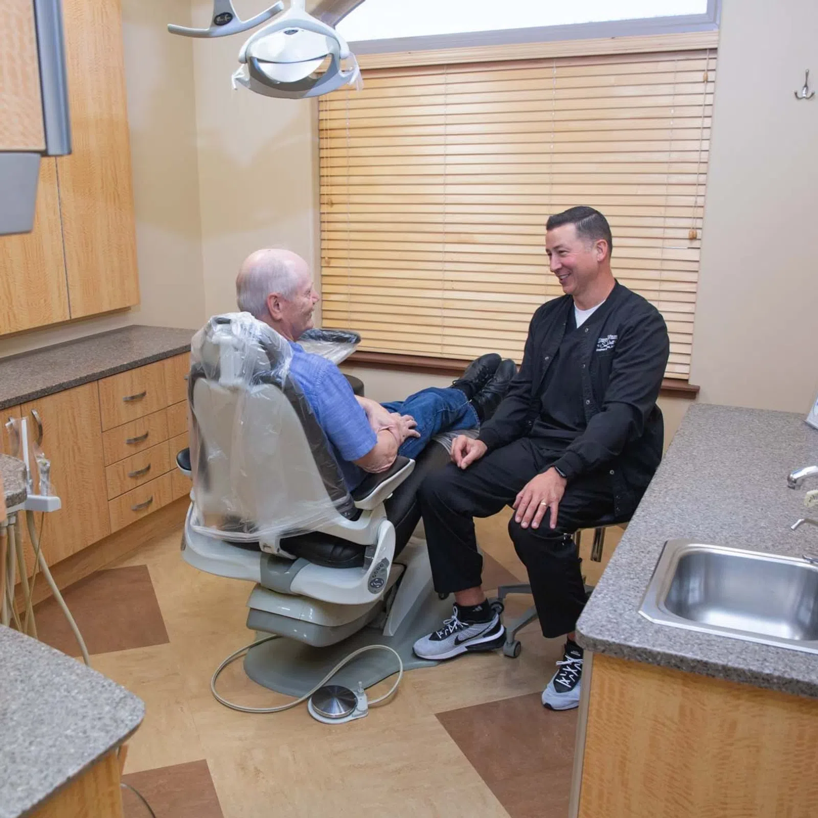 Dentist talking and smiling with elderly male patient seated in dental chair in modern clinic room.
