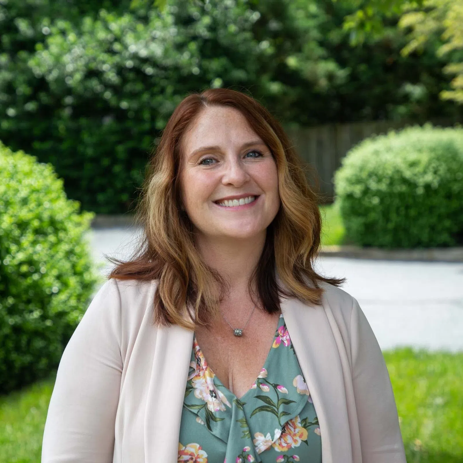 Smiling woman with brown hair in floral dress and beige blazer standing outdoors with green bushes in background