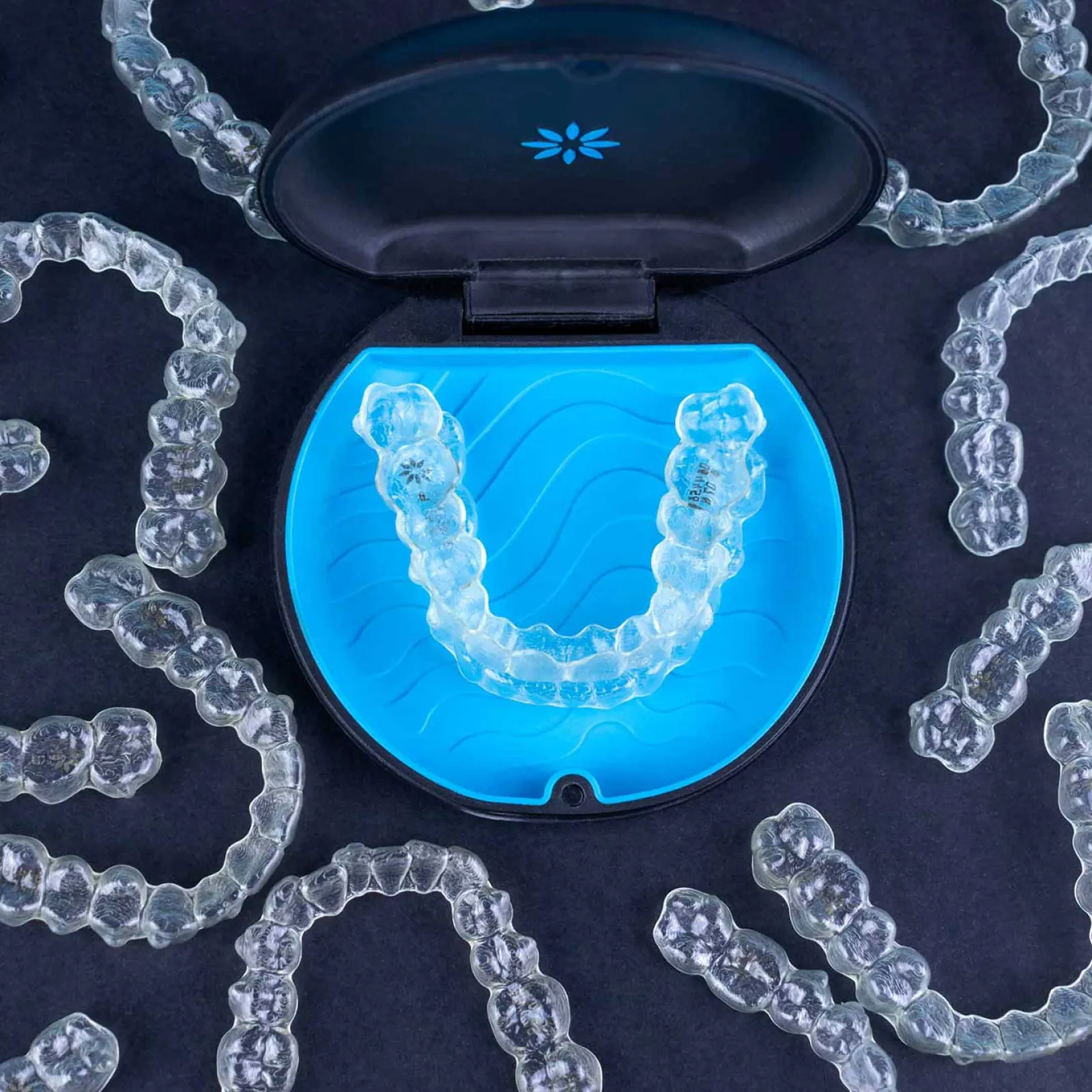 Clear dental aligners displayed around an open blue case on a dark background for teeth straightening.