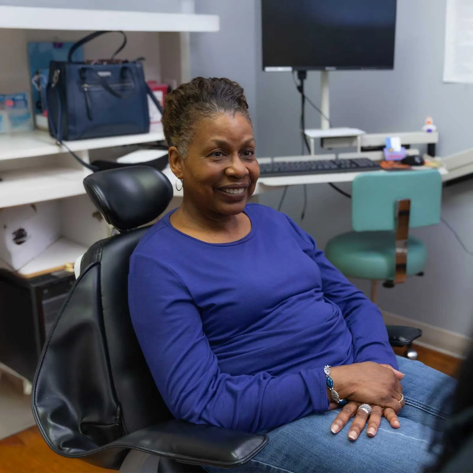 Smiling woman in blue shirt sitting in chair, talking to someone in an office with computer and shelves.
