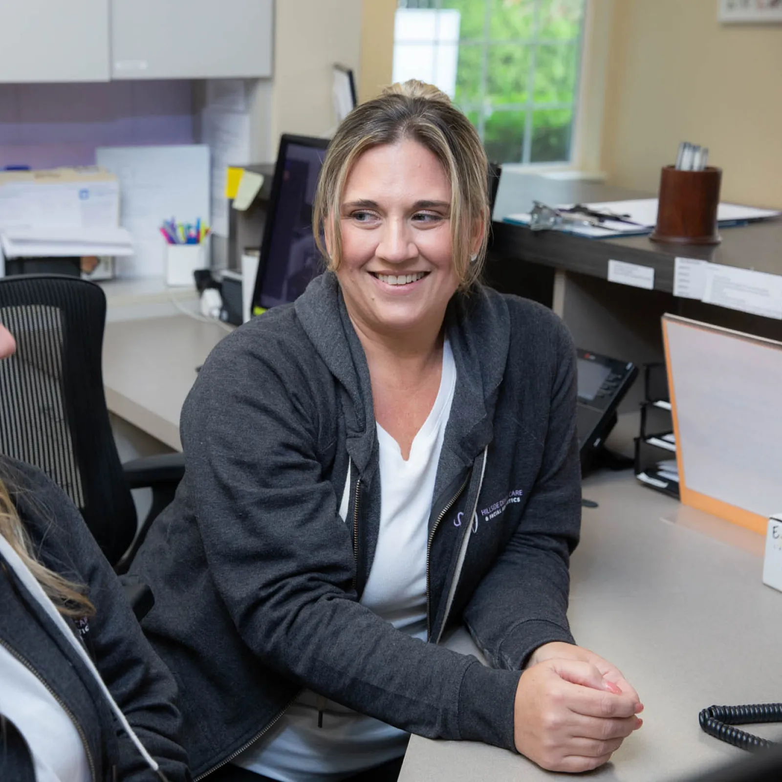 Two women in casual workwear smiling and conversing at an office desk with computer and supplies.