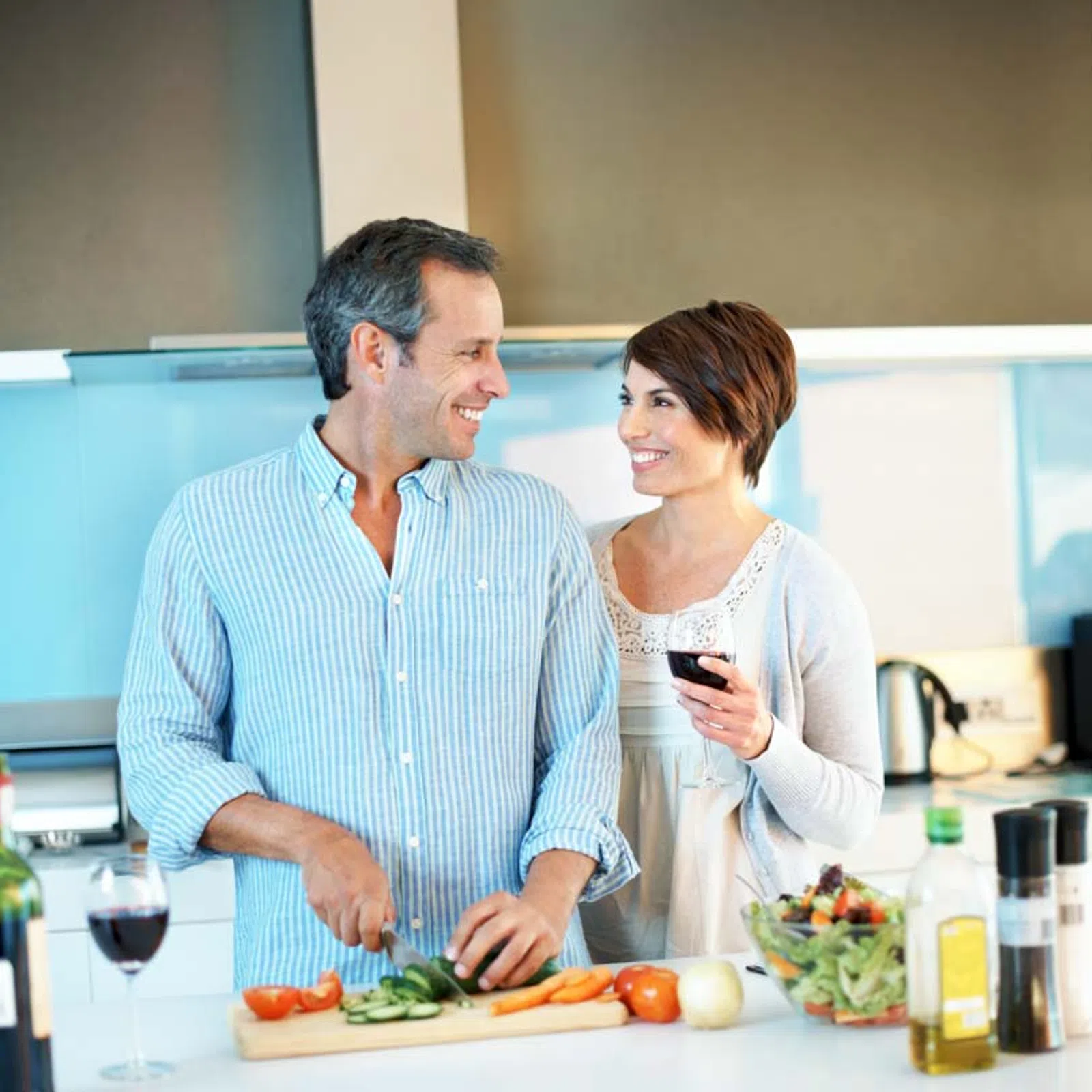 Couple smiling and preparing fresh vegetables in the kitchen while enjoying a glass of red wine together.