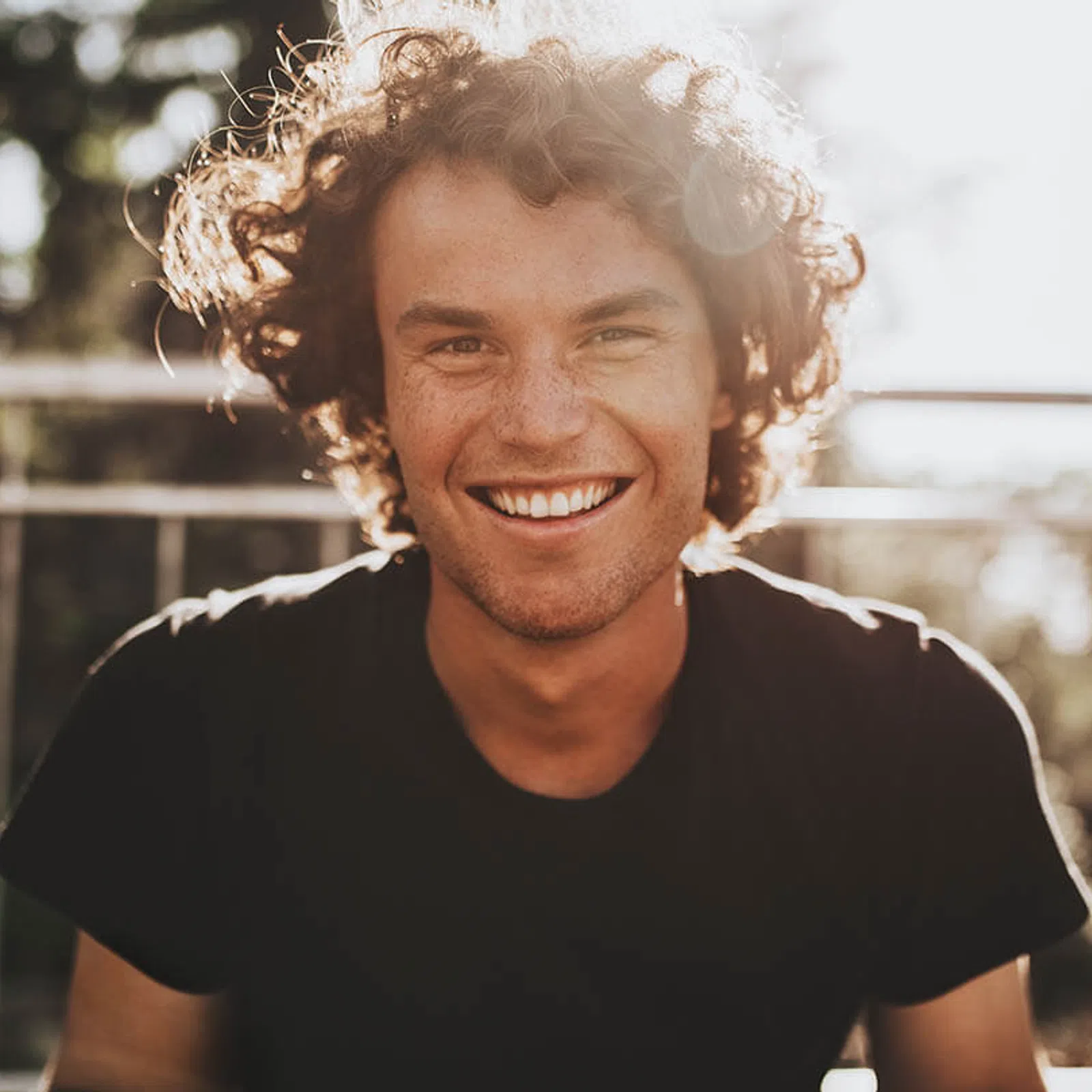 Young man with curly hair smiling brightly outdoors in sunlight wearing a black t-shirt.