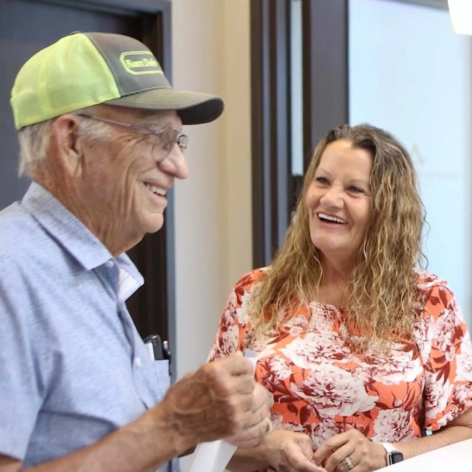 Elderly man in cap and woman in floral shirt smiling and chatting indoors at a reception desk.