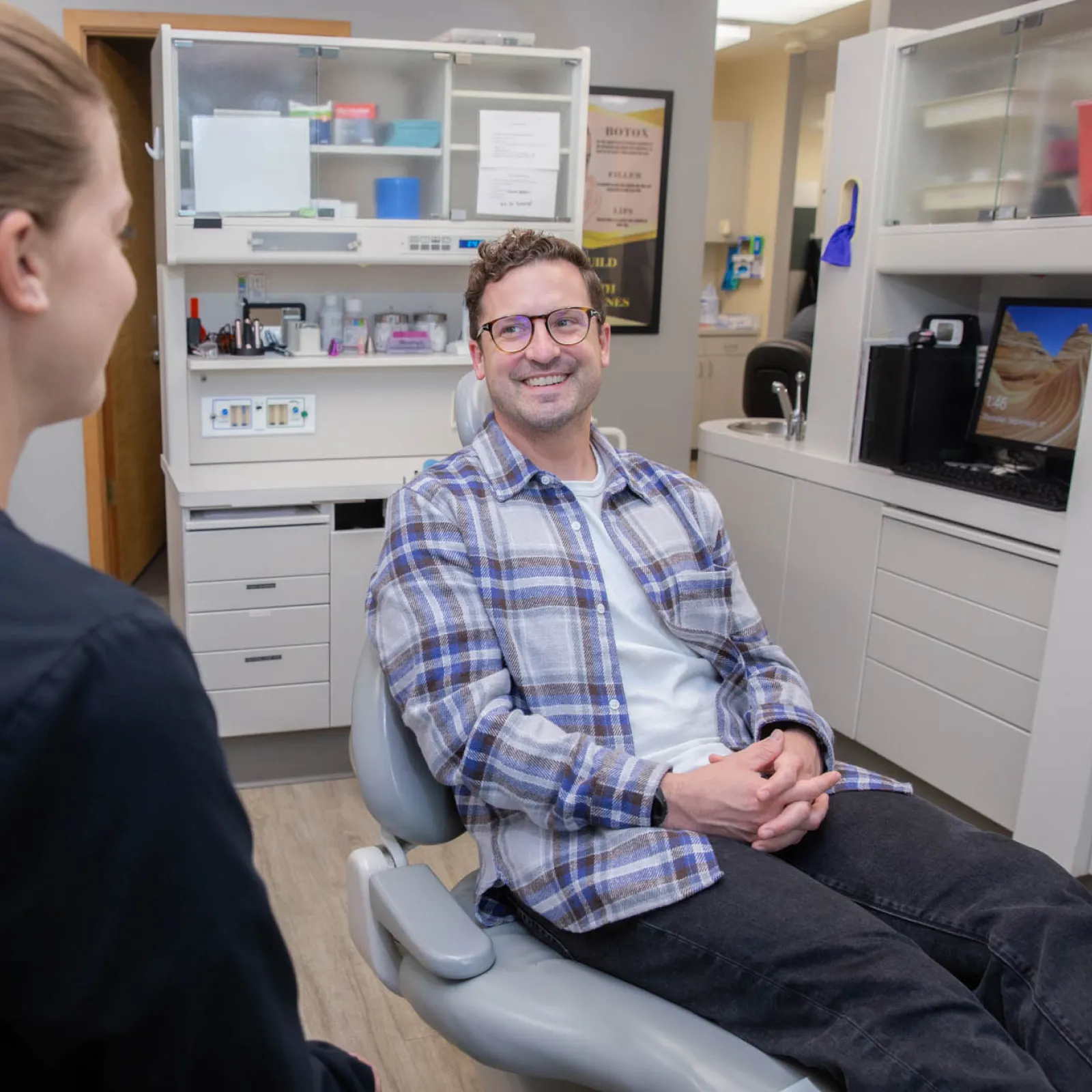Male patient smiling in dental chair while talking to female dentist in modern clinic room.