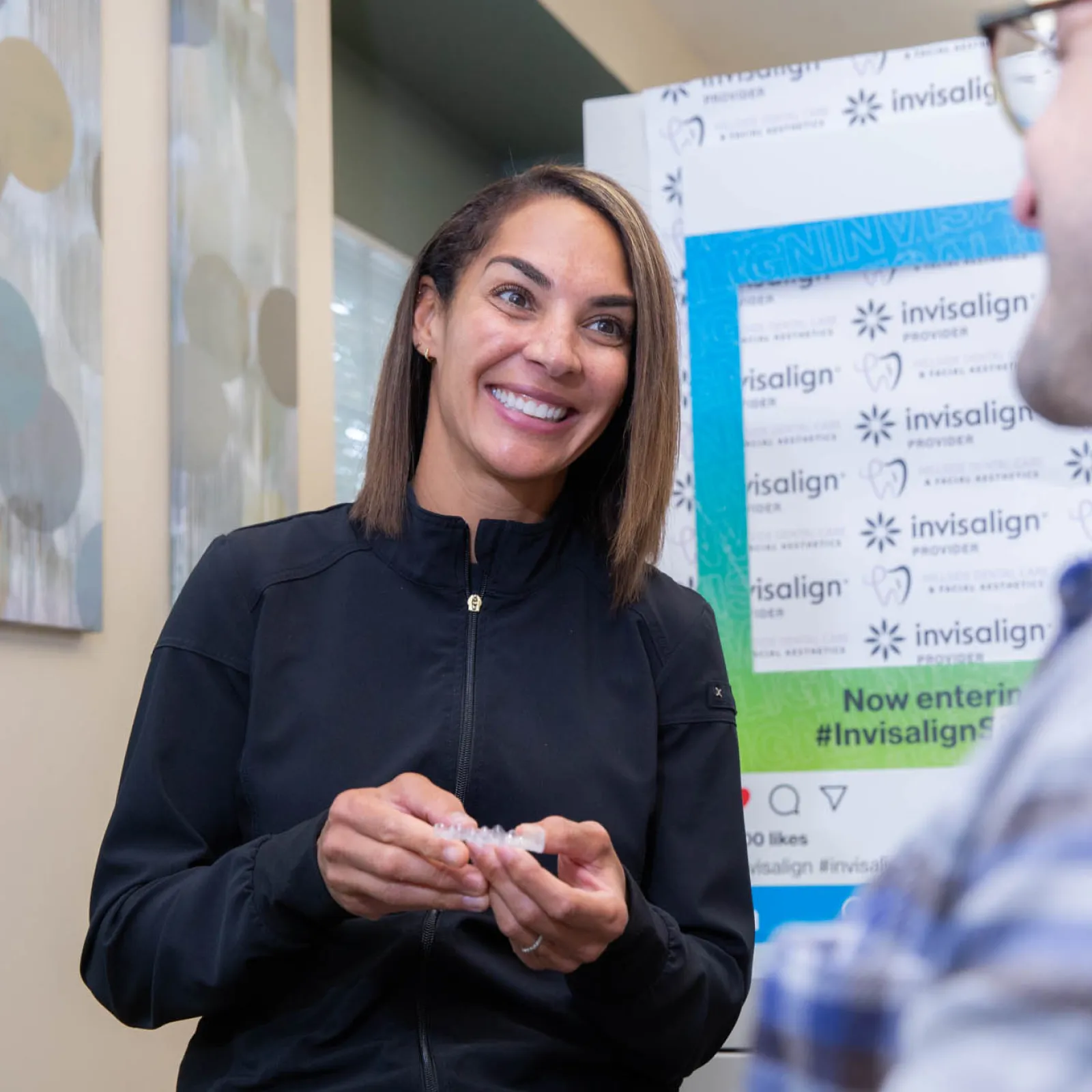 Smiling dental professional explaining Invisalign aligners to a male patient in a consultation room.