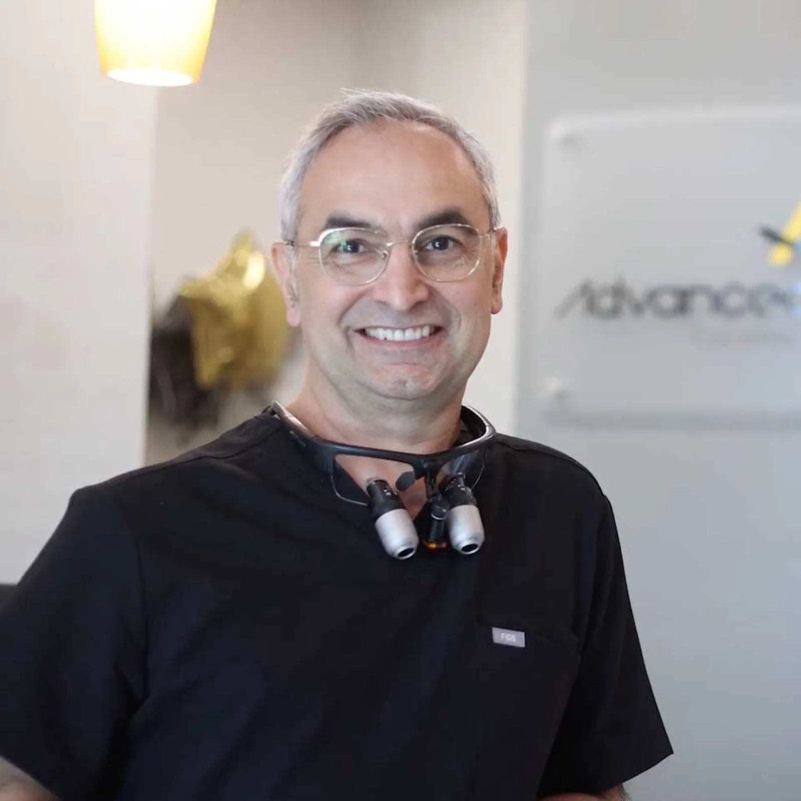 Smiling middle-aged male dentist in black scrubs standing in Advanced Dental Care reception area.