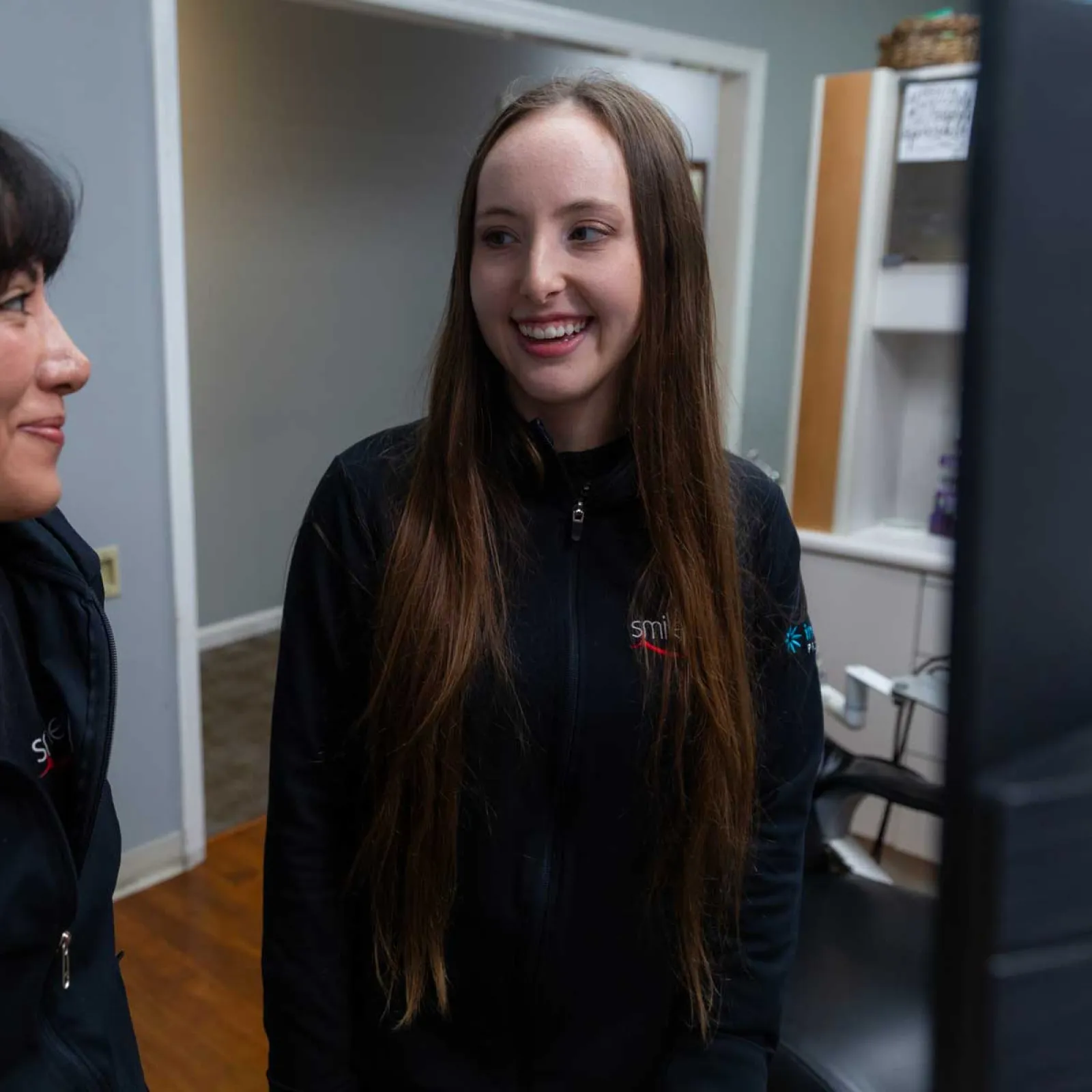 Two women in black jackets smiling and chatting in a modern office setting with wood flooring and shelves.