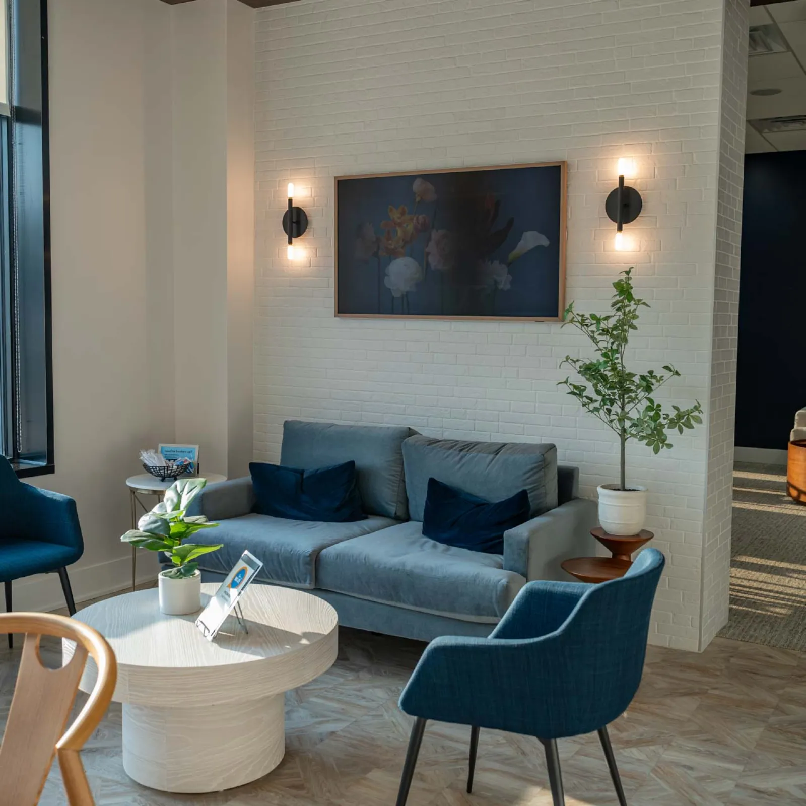 Bright modern waiting room with blue chairs, gray sofa, wooden table, and wall art on white brick wall.