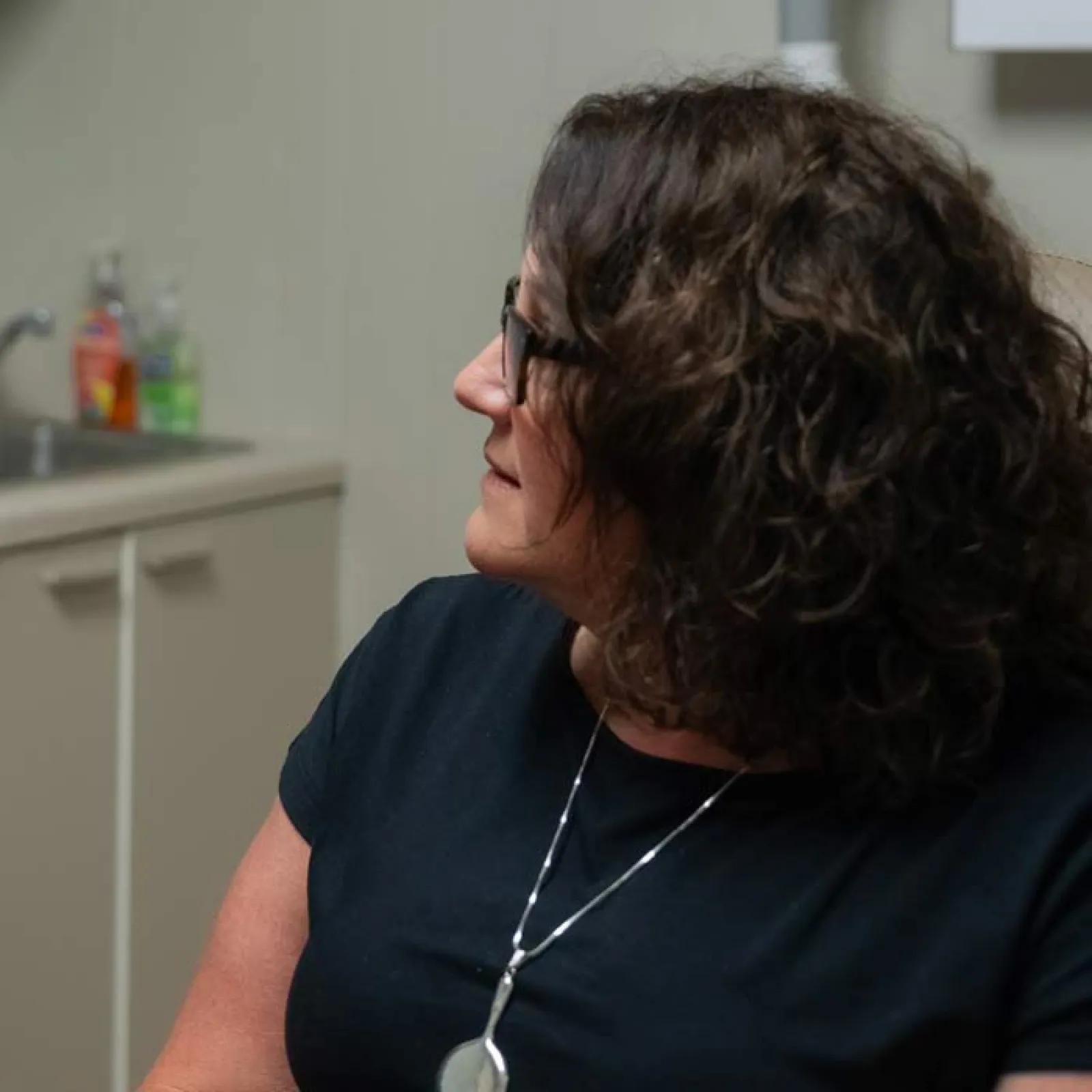 Woman with curly hair and glasses wearing a black shirt and silver necklace looking to the side indoors.