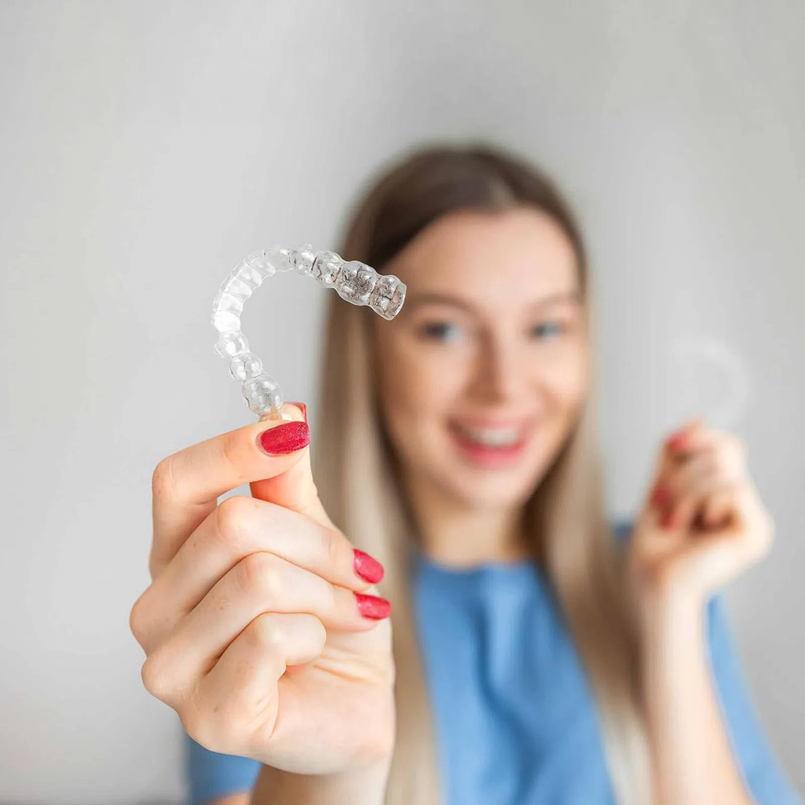 Smiling woman holding clear dental aligners for teeth straightening in a bright, neutral setting.