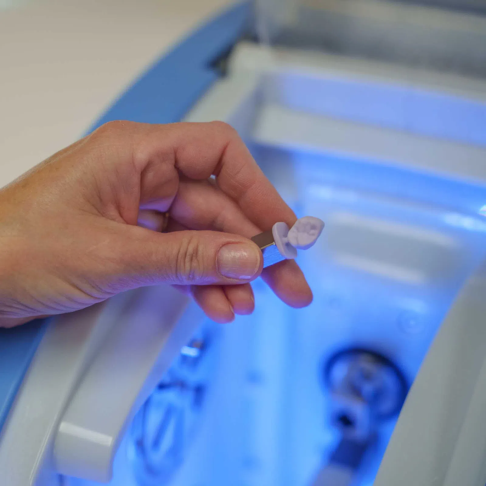 Hand holding a small lab sample tube near a blue-lit laboratory machine for testing or analysis.