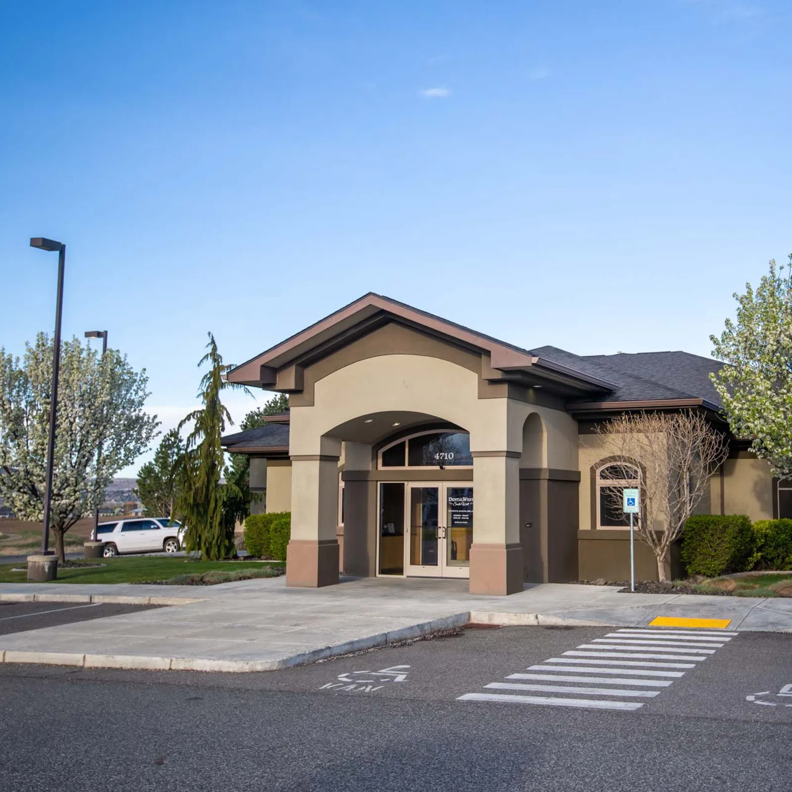 Modern beige single-story office building with arched entrance surrounded by blooming trees and parking spaces.