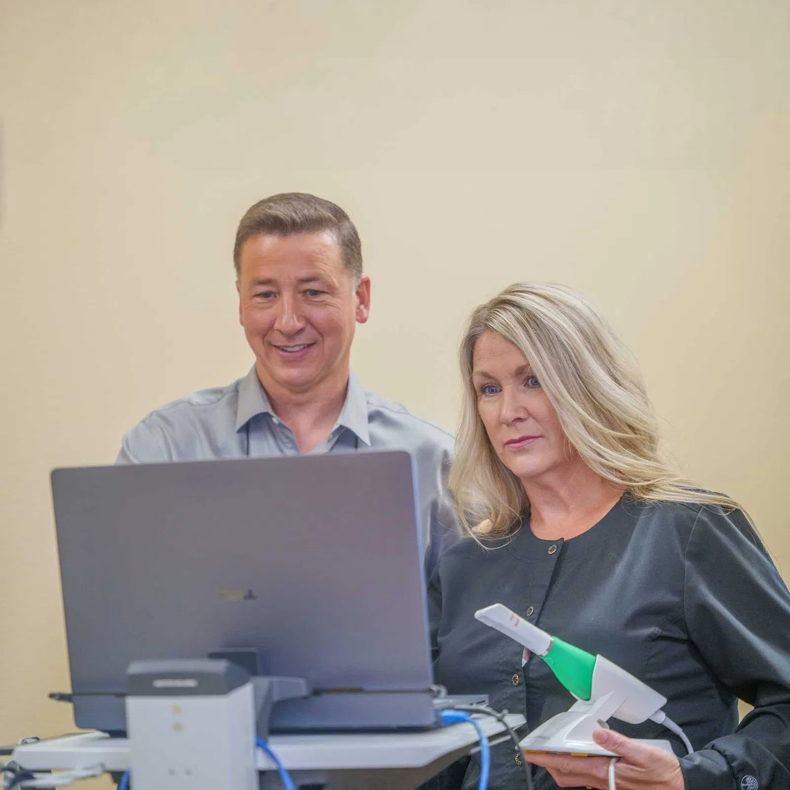 Two professionals reviewing data on a laptop with a handheld scanner in a neutral office setting