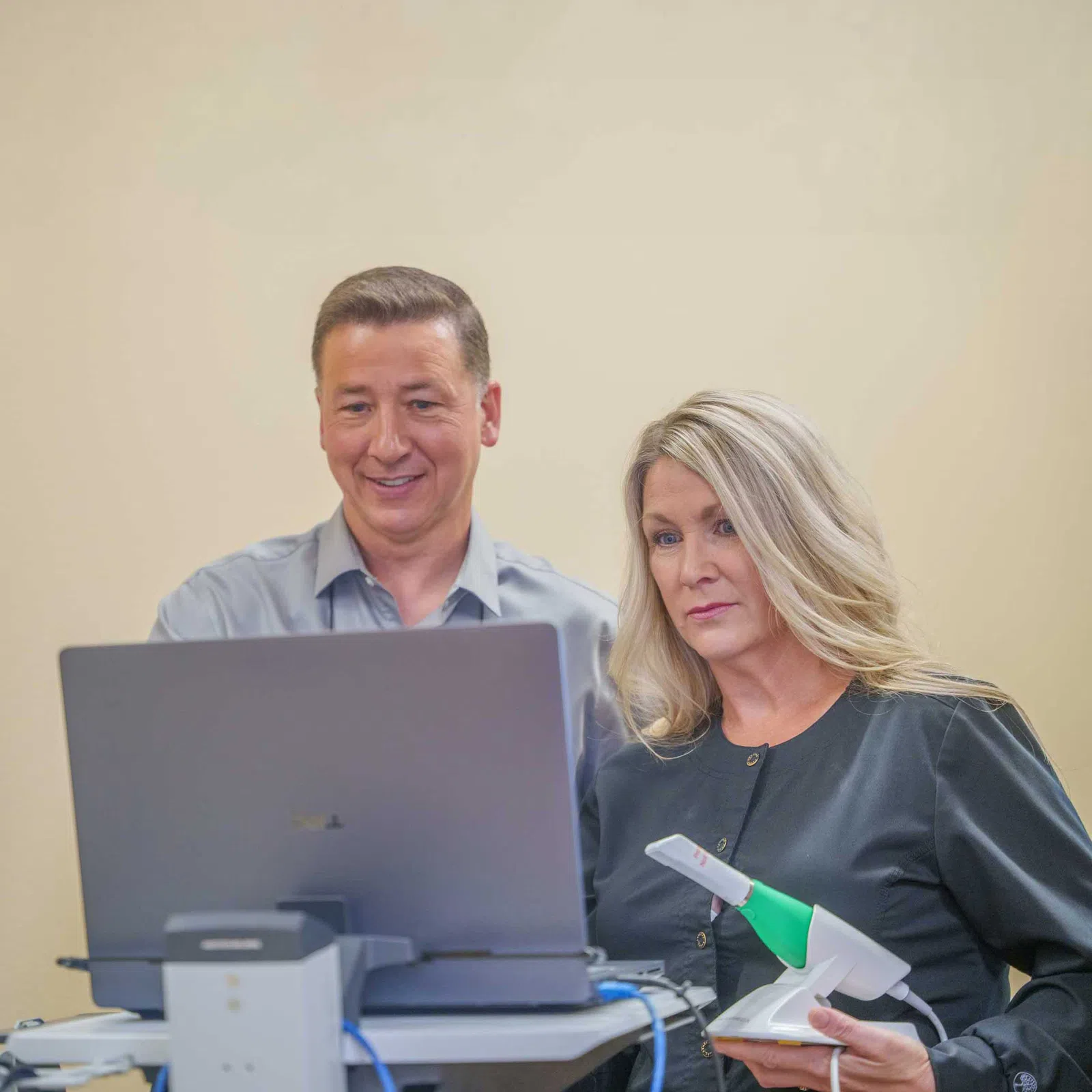 Two professionals working together at a laptop, one holding a handheld device with a green and white tip.