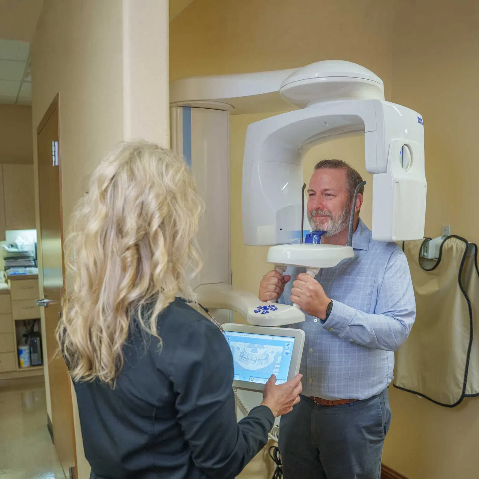 Man undergoing dental 3D X-ray scan operated by a female technician in a dental office with beige walls.