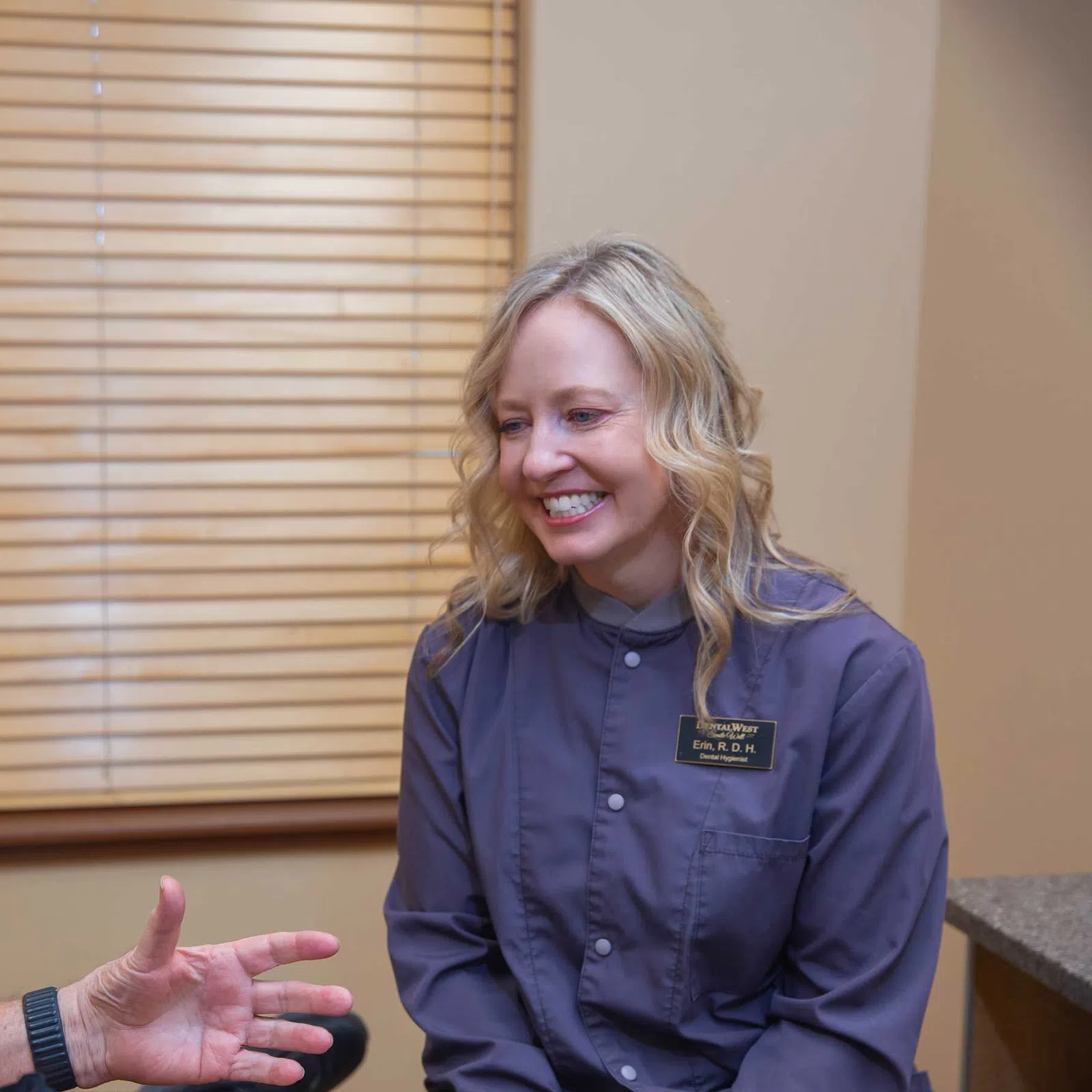 Smiling female healthcare professional consulting an elderly male patient in a clinic room with wooden blinds.