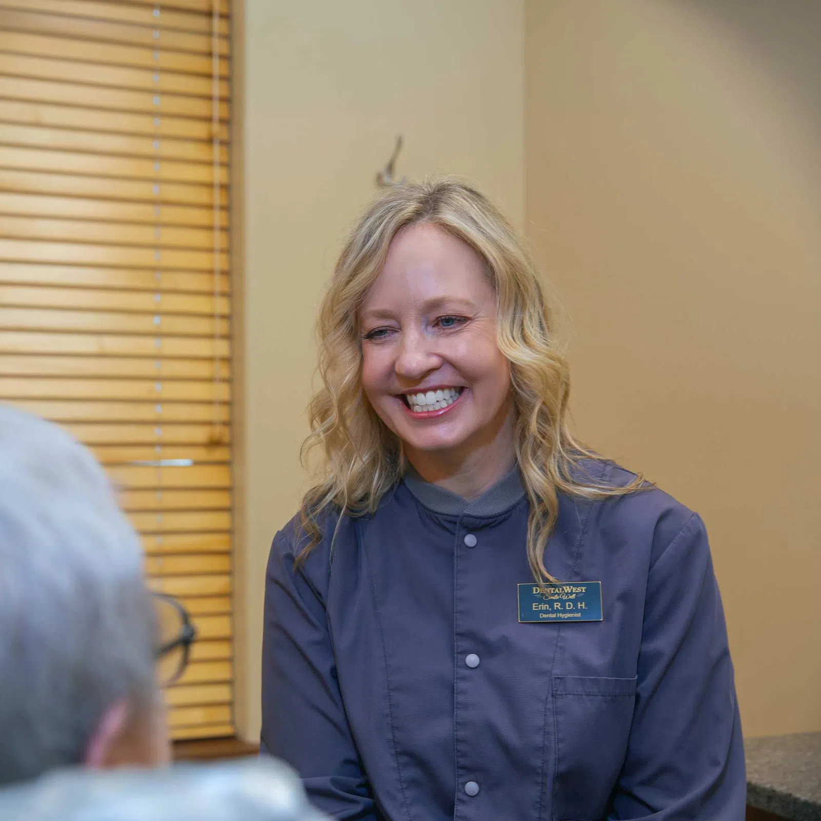 Smiling female healthcare professional talking to elderly patient in a warmly lit office with wooden blinds.