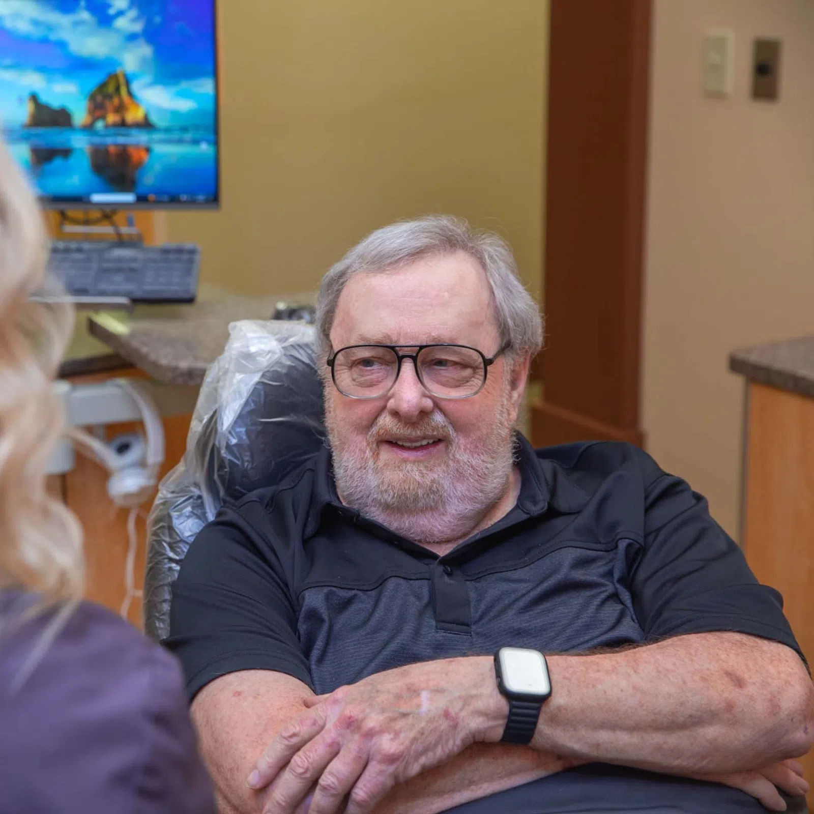 Elderly man wearing glasses talking with a female healthcare professional in a medical office setting.
