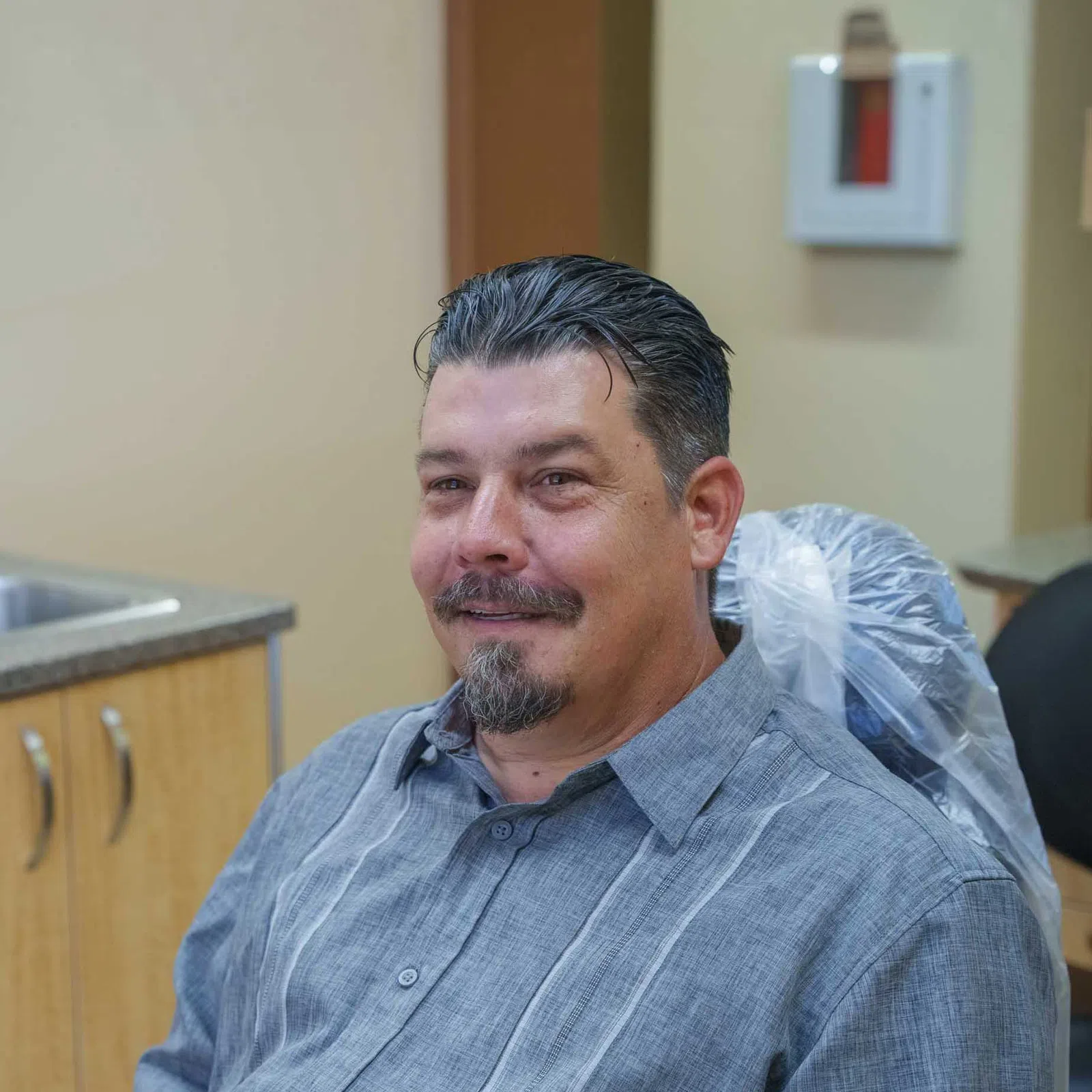 Smiling man with slicked-back hair and goatee seated in a dental office chair with protective cover.