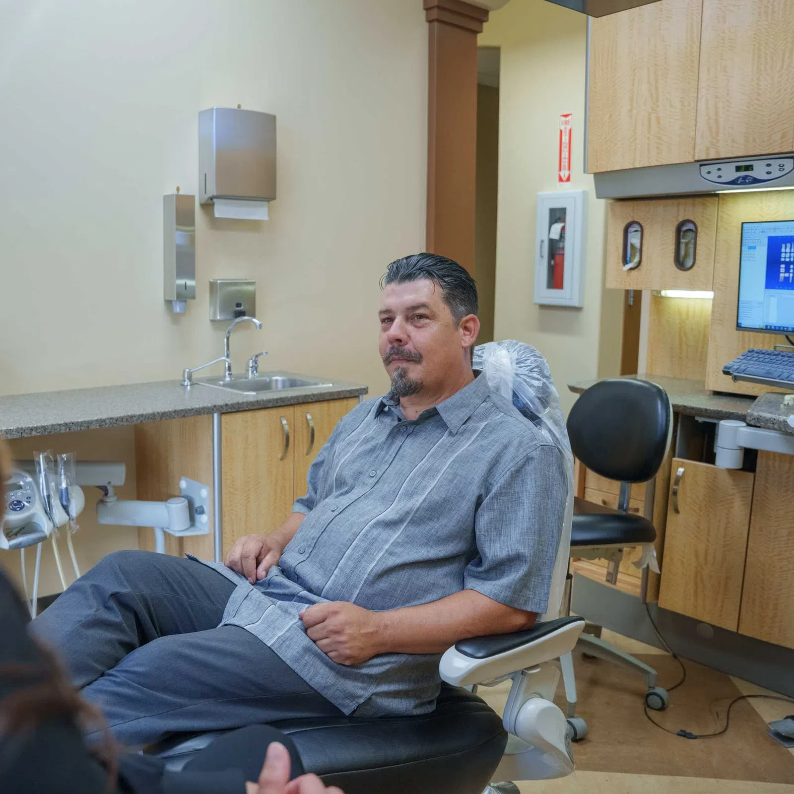 Man sitting in a dental chair facing a woman in a clinic with dental equipment and computer screens visible.
