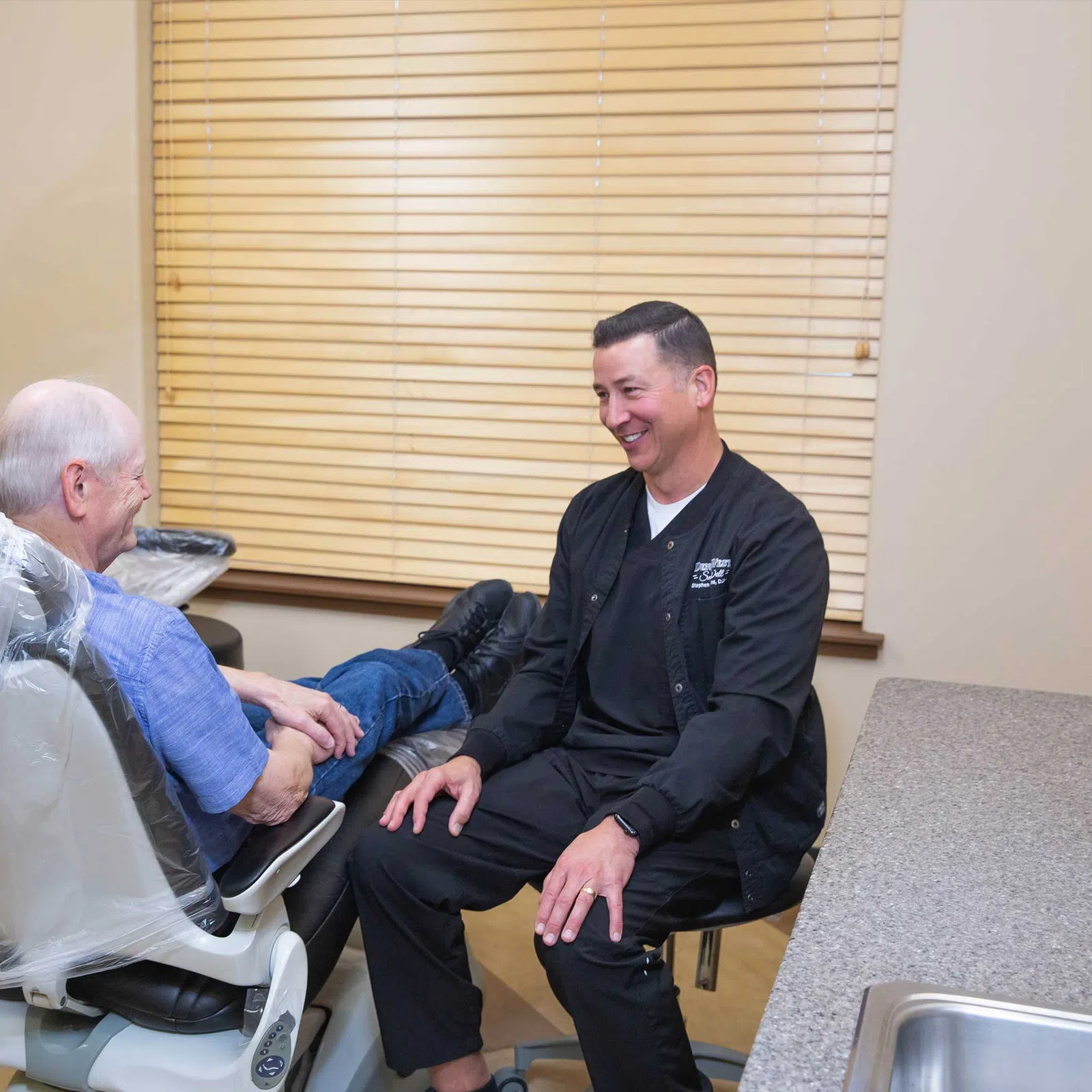 Dentist in black uniform smiling and talking with elderly male patient sitting in dental chair in clinic.