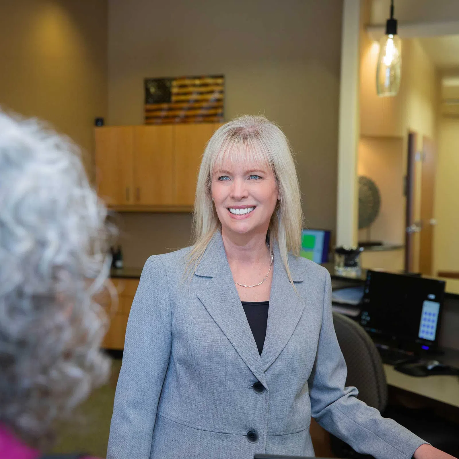 Professional woman in gray blazer smiling and interacting with a client in an office setting.