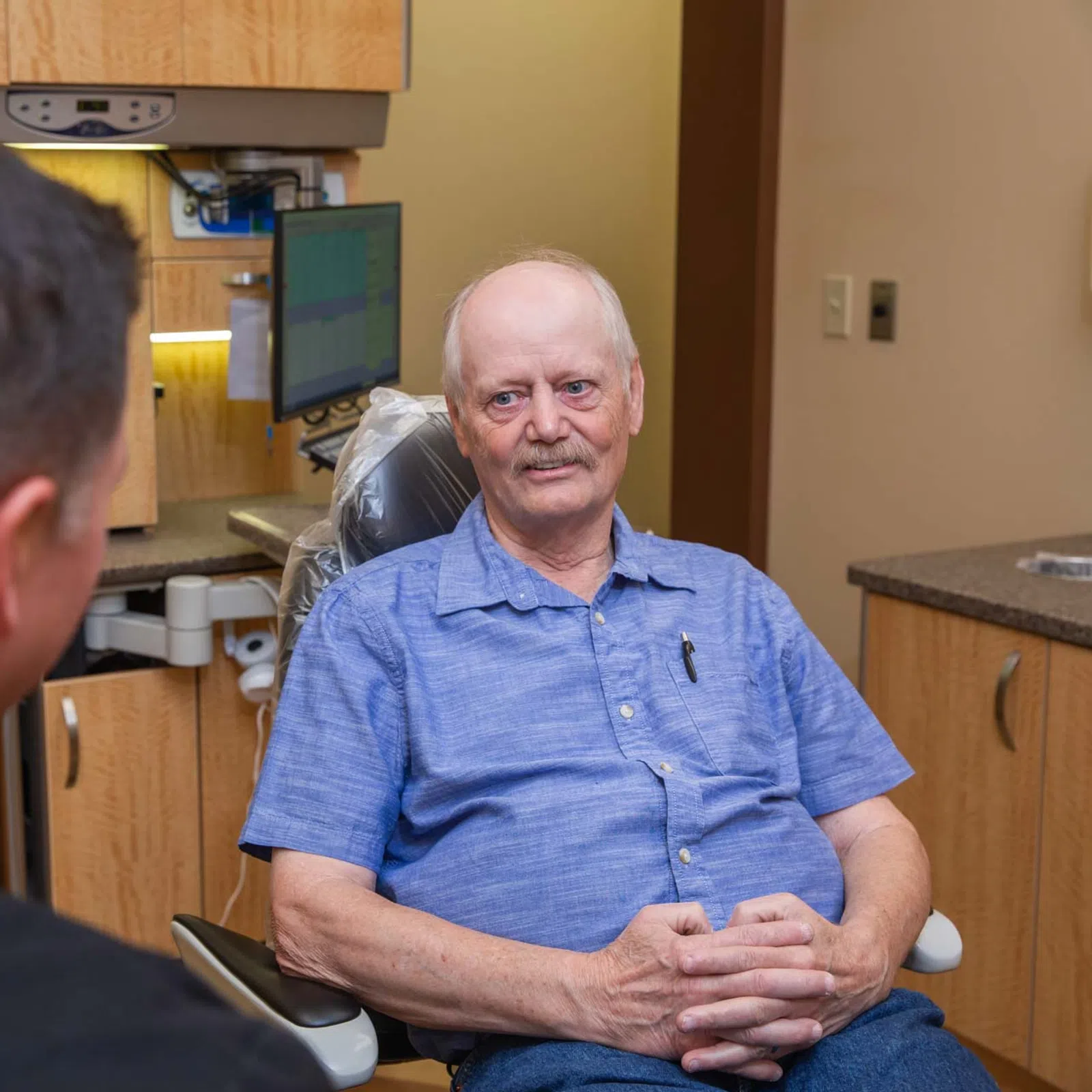 Elderly man in blue shirt sitting in a medical office chair talking to a healthcare professional.