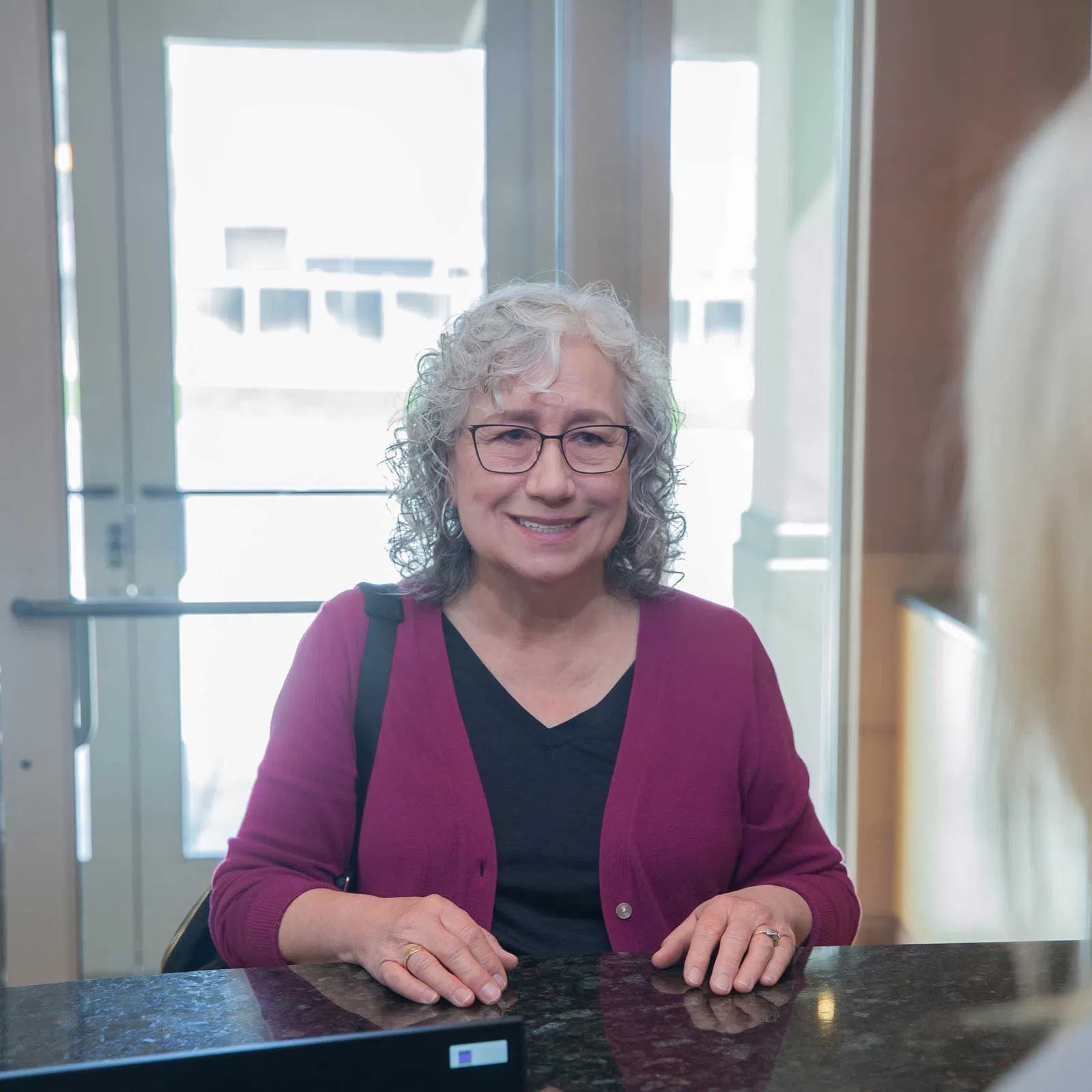Smiling mature woman with glasses and curly gray hair wearing a maroon cardigan at reception desk.