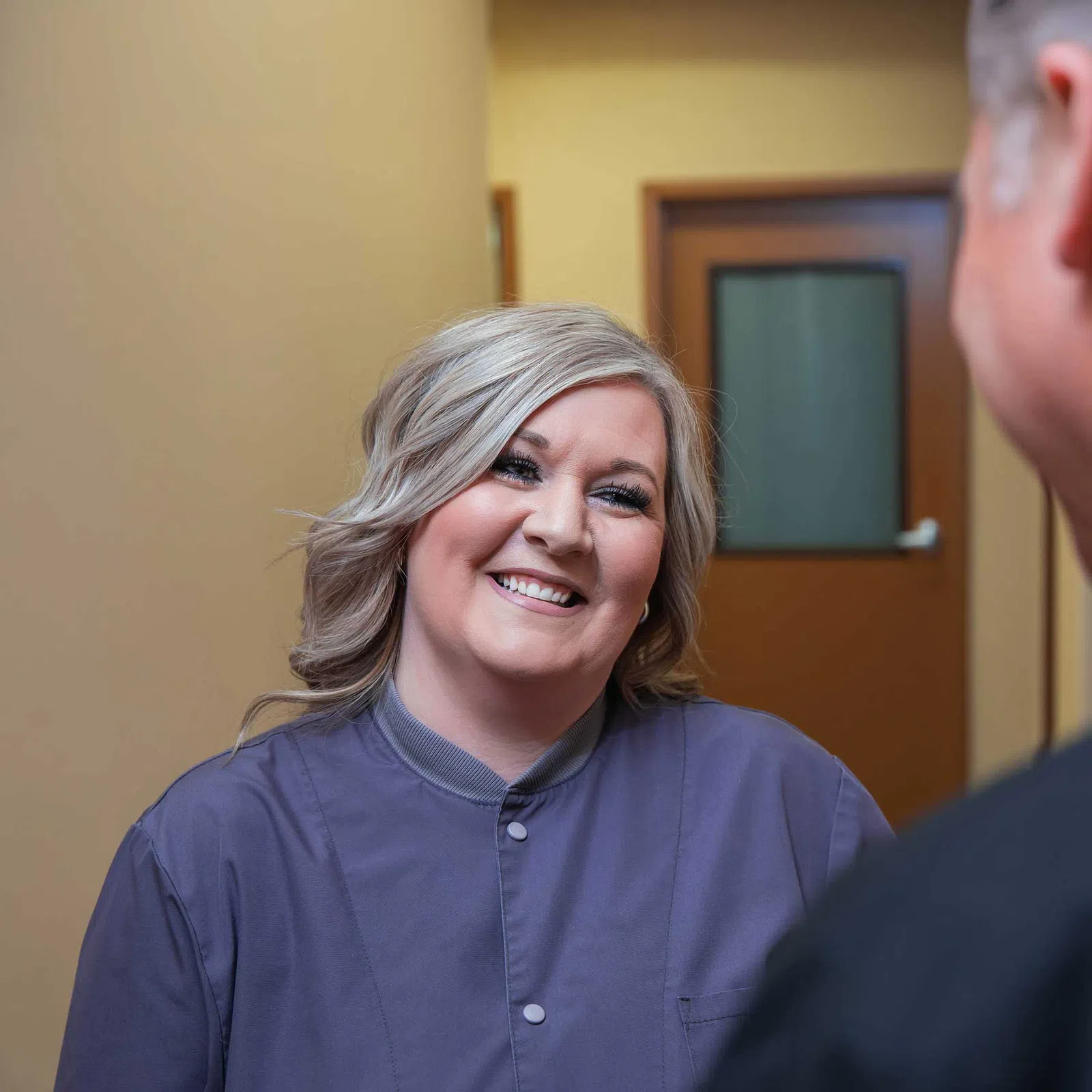 Smiling woman in purple scrubs talking to a man in a hallway with a door in the background