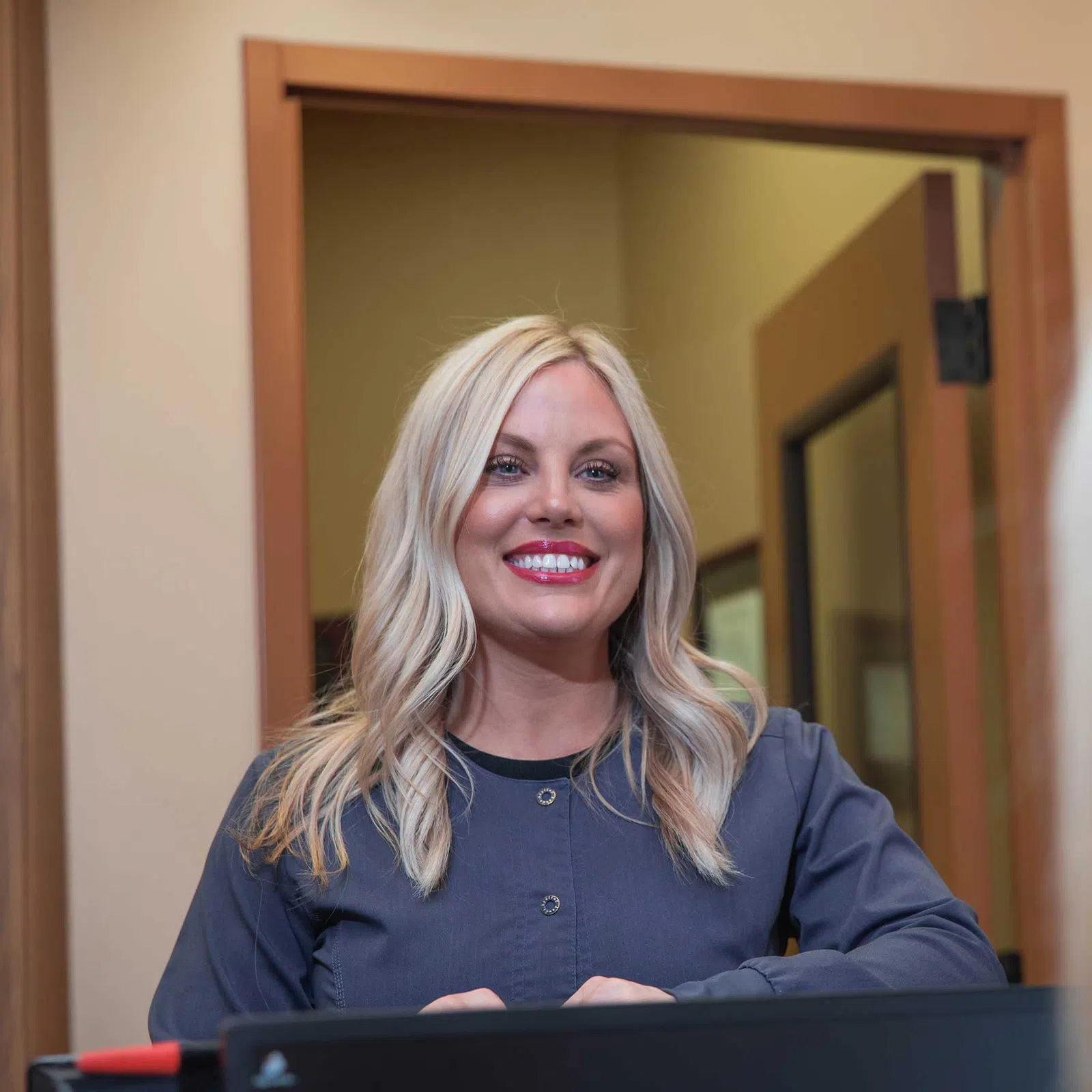 Smiling blonde woman with long hair wearing navy blue scrubs stands behind reception desk in office.