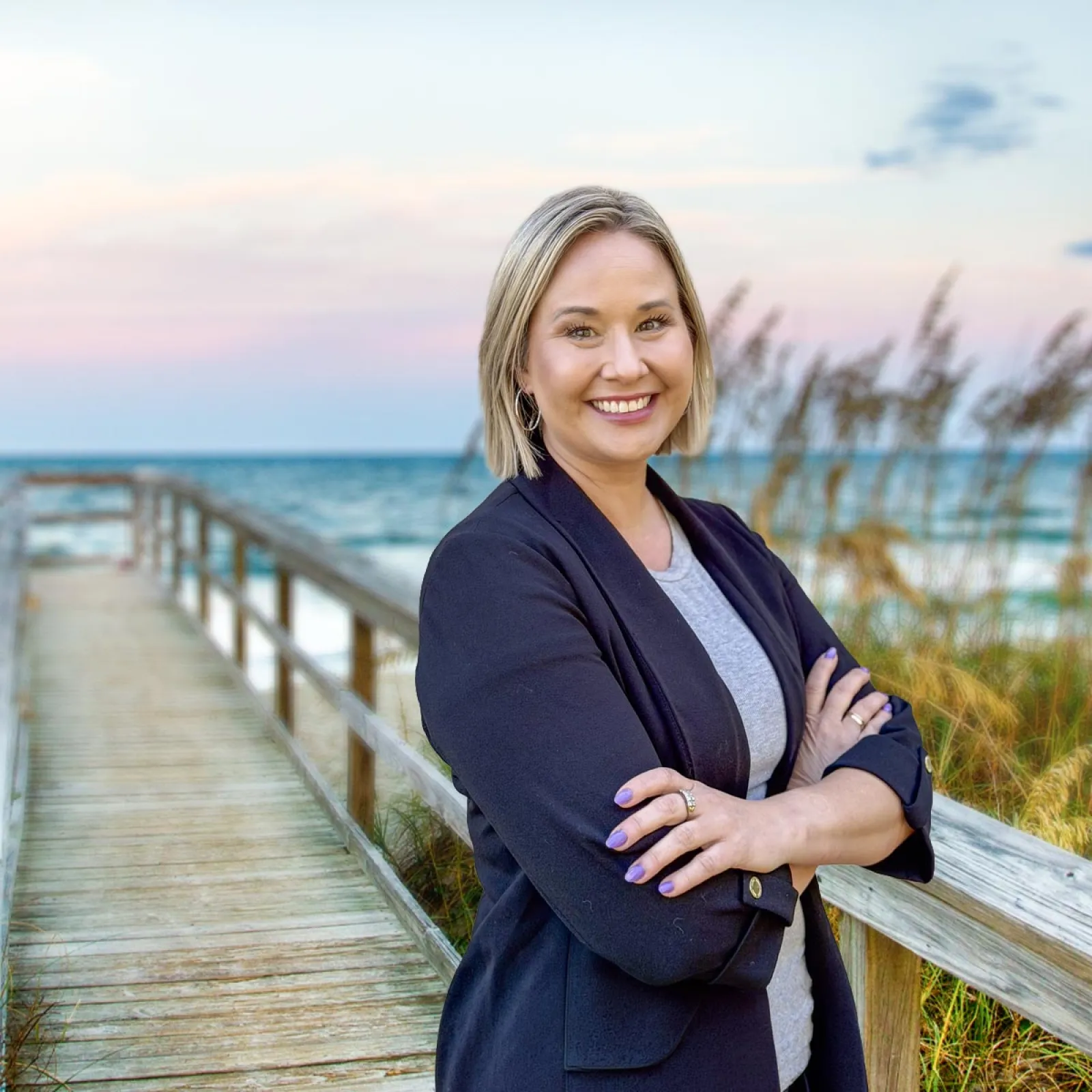 Smiling woman with crossed arms standing on wooden boardwalk by beach during sunset.
