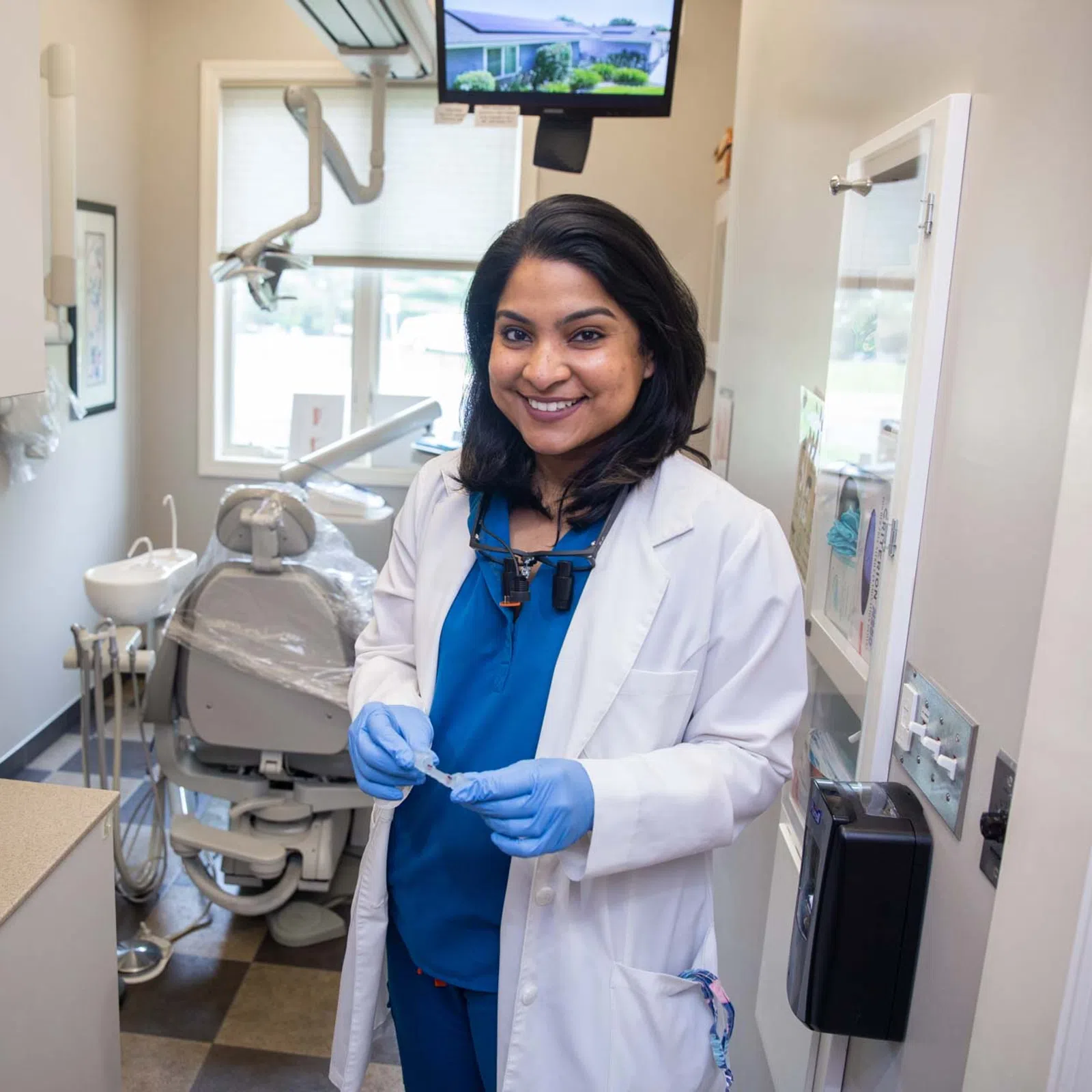 Smiling female dentist wearing blue scrubs and white coat in a modern dental office with equipment