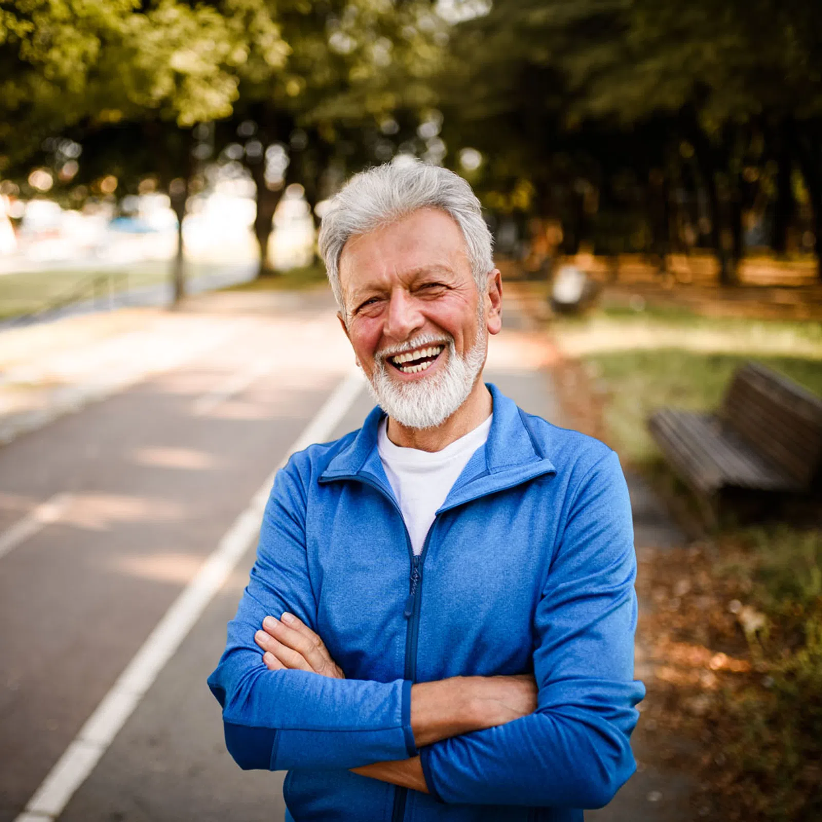 Smiling senior man in blue jacket standing with arms crossed on park path with trees and benches.