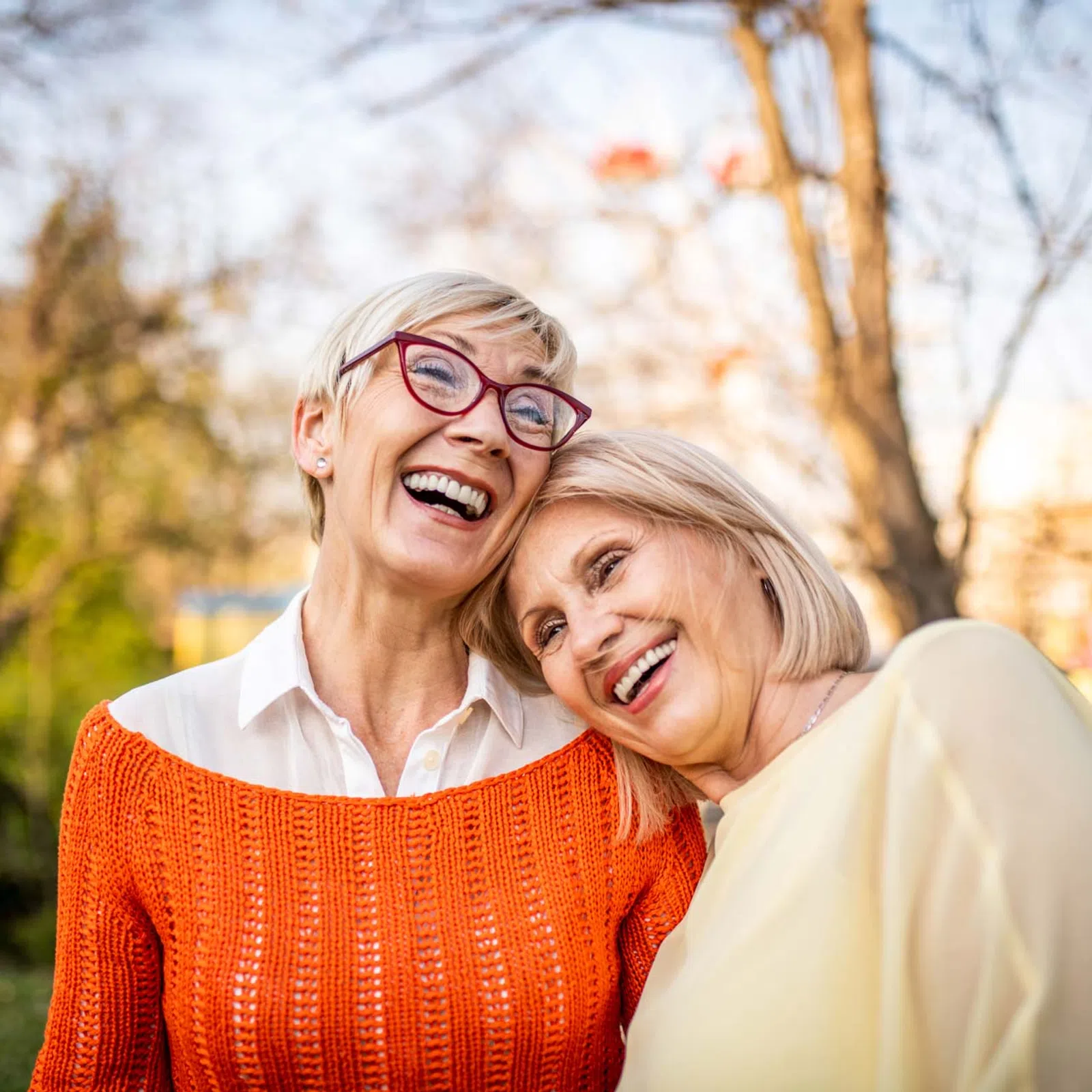 Two happy mature women smiling outdoors, one wearing an orange sweater and red glasses, the other in a light blouse.