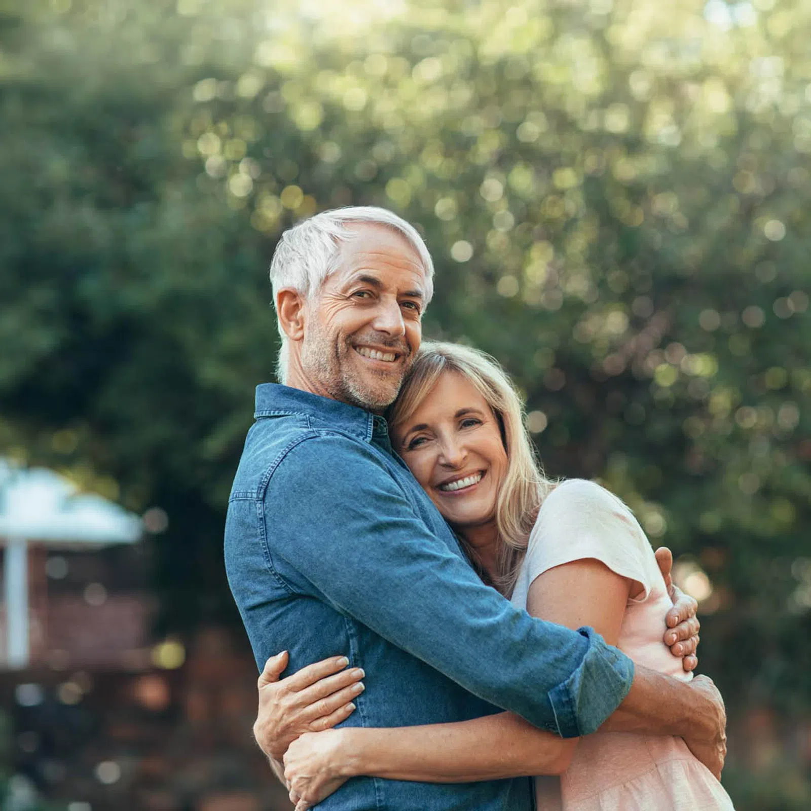 Smiling mature couple hugging outdoors with greenery and house in the background on a sunny day