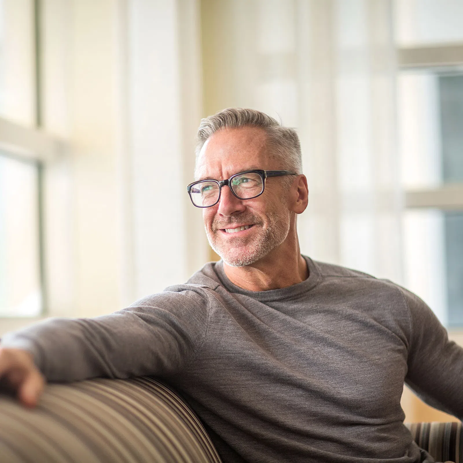 Smiling middle-aged man with gray hair and glasses sitting on a striped couch in a bright living room.