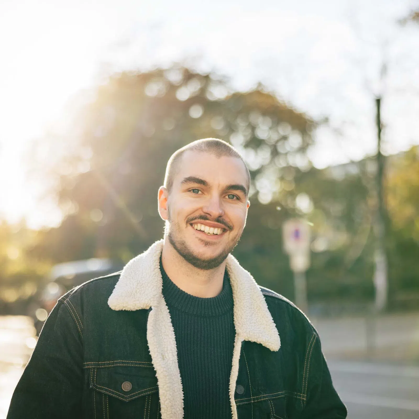 Smiling young man wearing a denim jacket with shearling collar outdoors on a sunny autumn day.