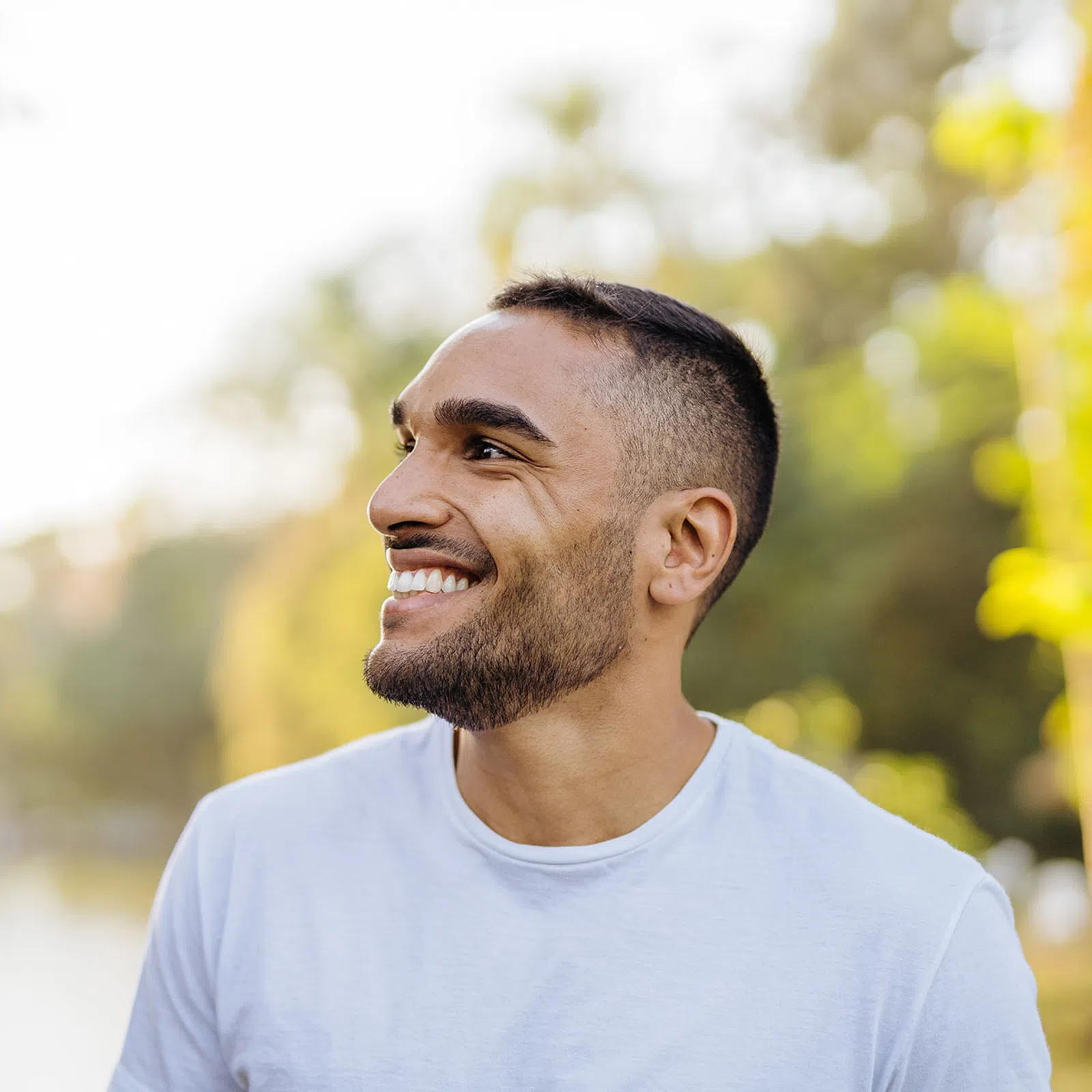 Smiling young man with beard wearing white shirt outdoors near blurred greenery on sunny day.