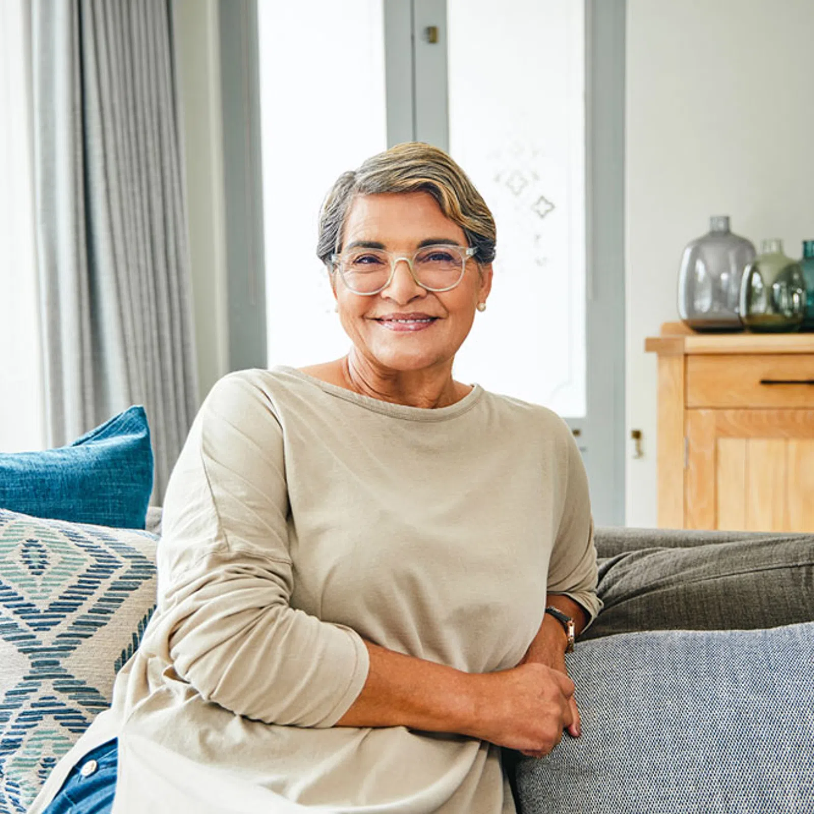 Smiling middle-aged woman wearing glasses and beige top sitting on a couch with decorative pillows in a bright living room.